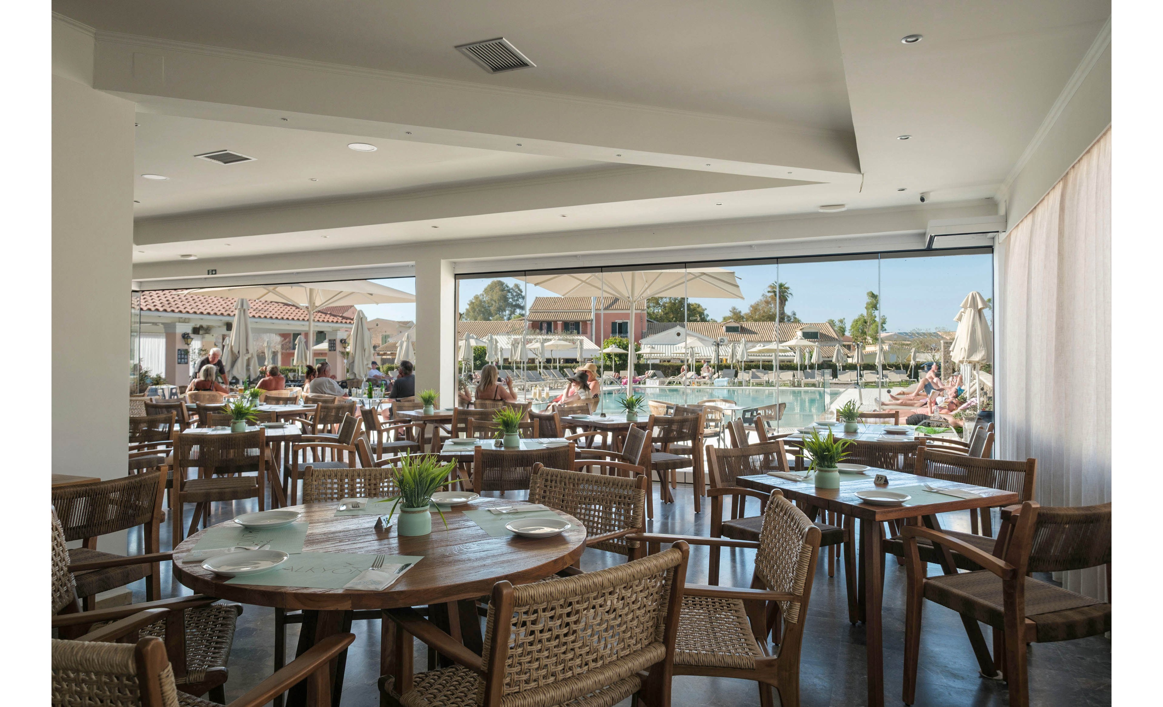 A bright open-air restaurant features wooden tables and woven chairs arranged neatly in a spacious interior. Small green plants decorate the tabletops, adding a fresh touch to the neutral decor. Large windows open out to a sunny pool area with loungers, umbrellas, and relaxed guests. The scene suggests a casual vacation dining experience at a resort or hotel.