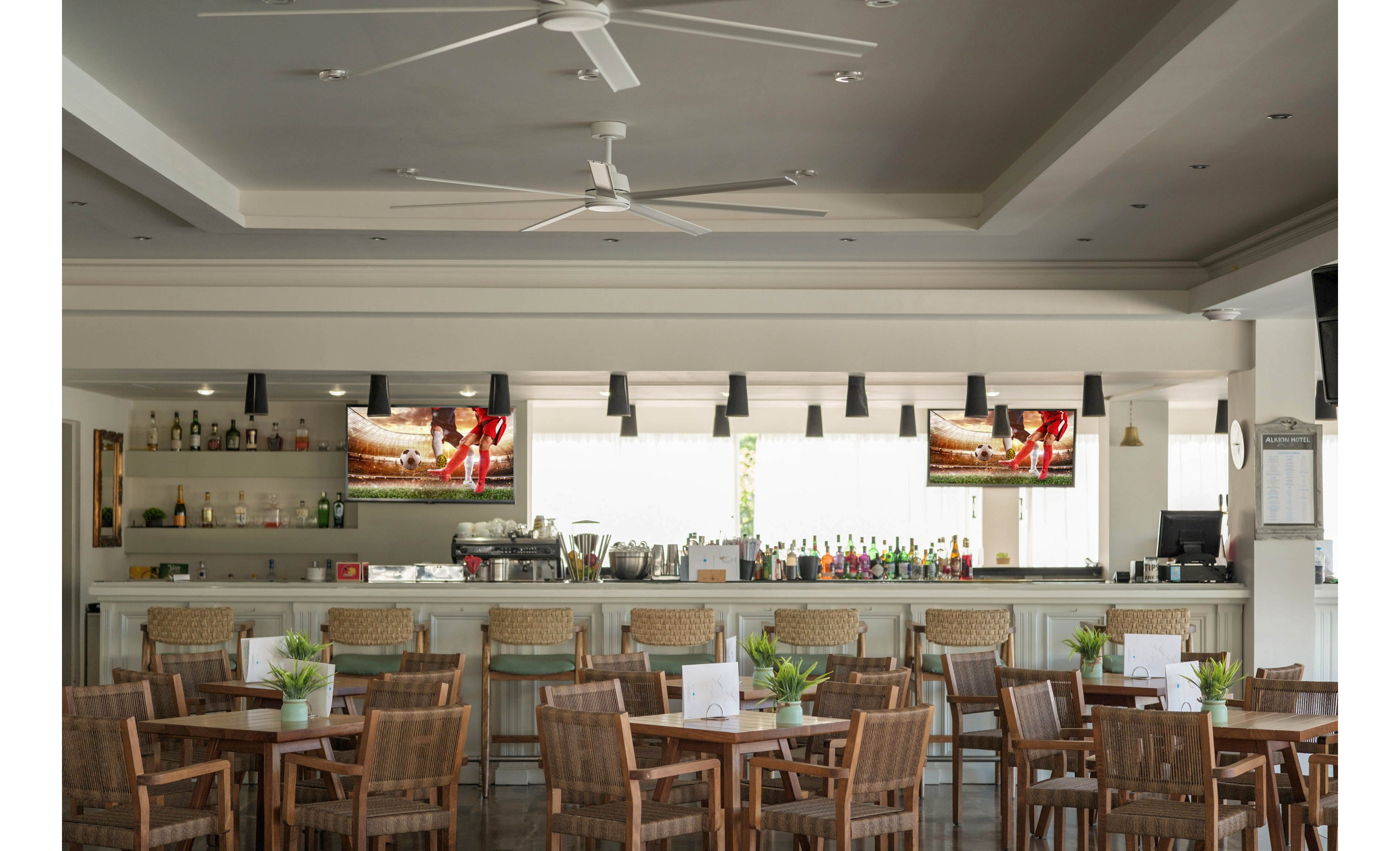 A contemporary indoor bar area features rows of empty wooden tables and chairs facing a long counter. Bottles and glassware are neatly arranged behind the bar, with two flat-screen televisions mounted above displaying a sports scene. Pendant lights hang from the ceiling alongside large white ceiling fans, creating a bright and airy atmosphere. Small potted plants and table menus add decorative touches to the relaxed, casual setting.