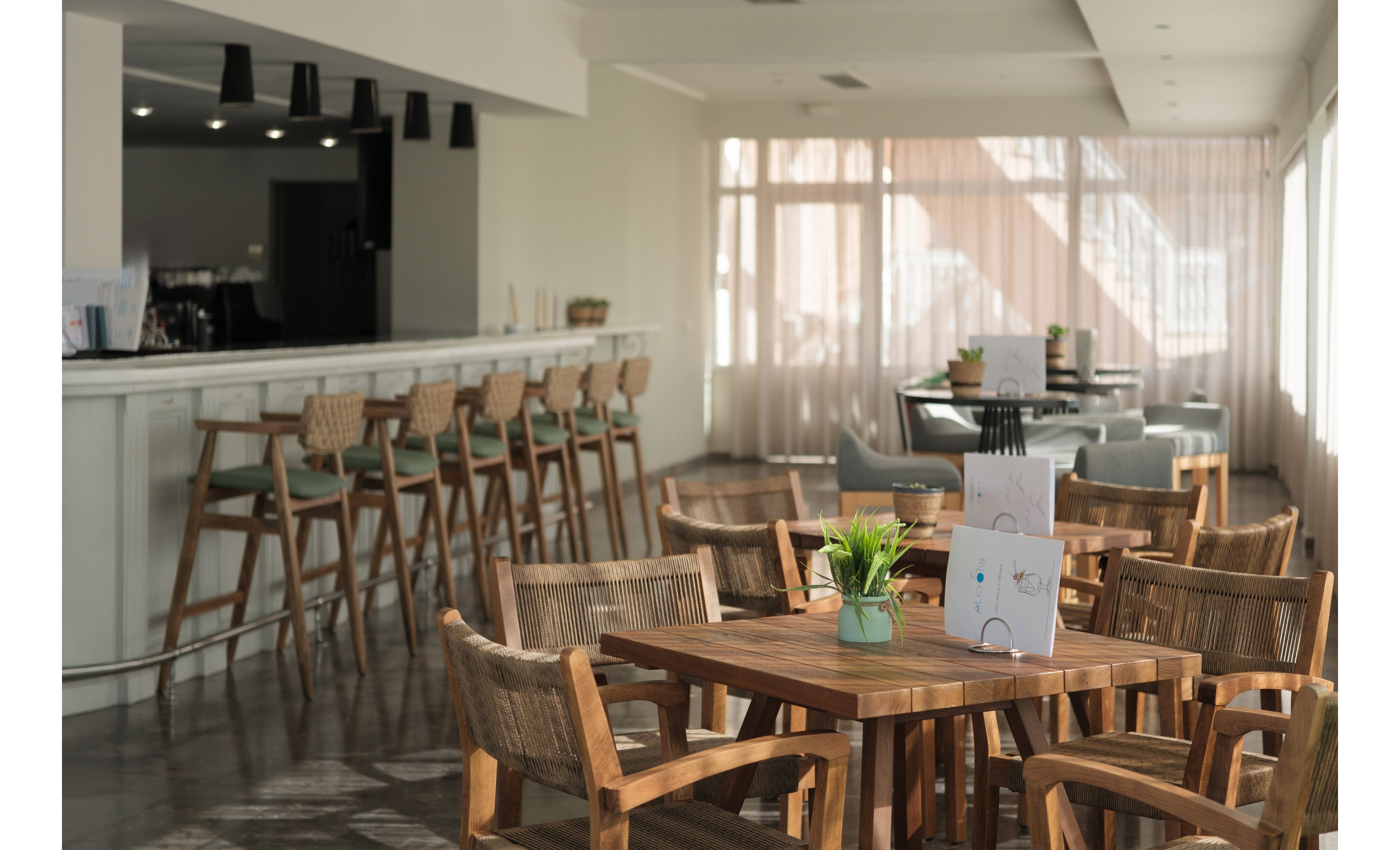 A bright, modern cafe interior features wooden tables and woven chairs in the foreground. Sunlight streams through large windows with sheer curtains, casting soft shadows across the polished floor. A long bar with high stools lines the left side, while additional seating is visible in the background. A small potted plant and table tent add detail to the central table, creating a relaxed and welcoming atmosphere.