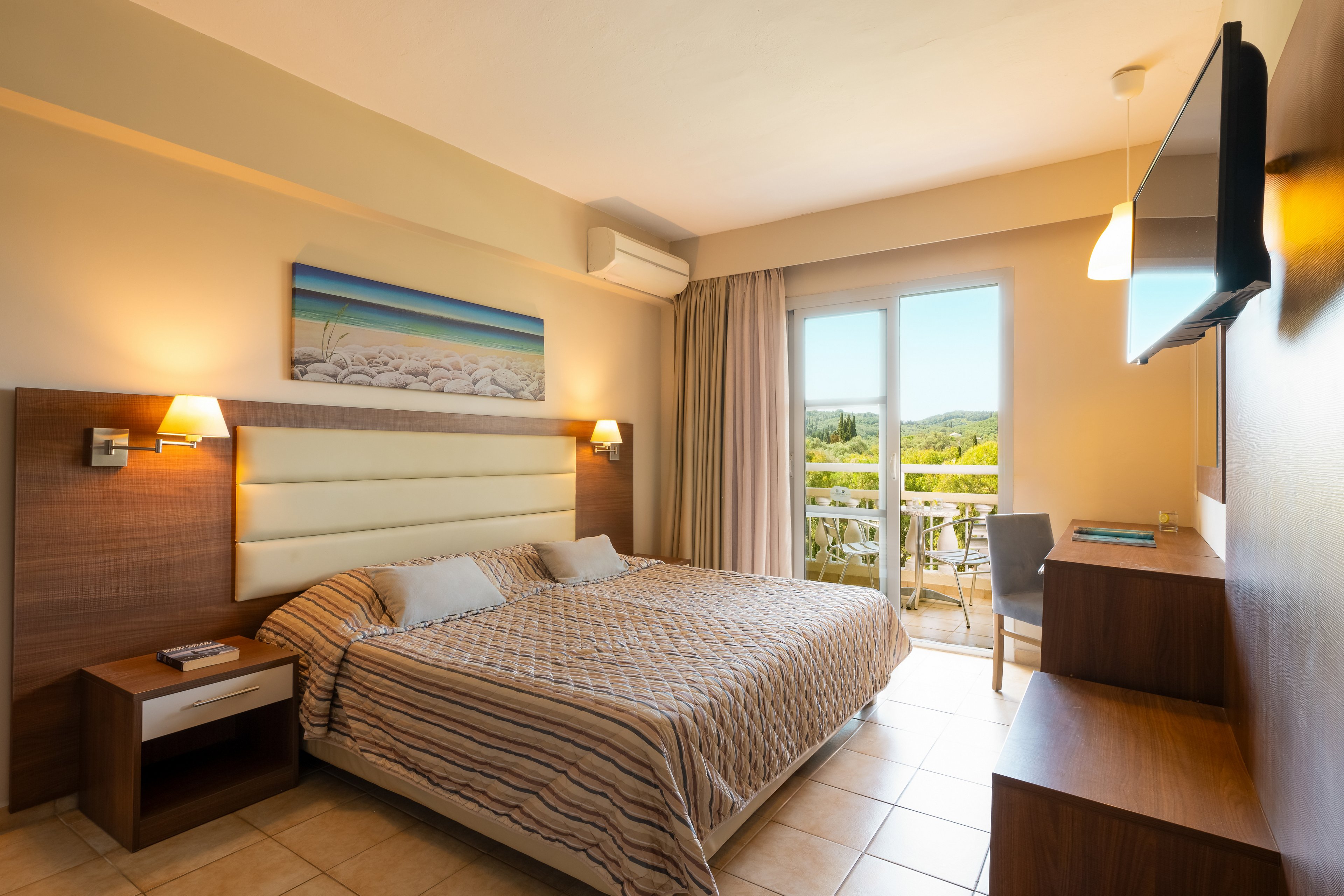 A well-lit hotel room featuring a double bed with striped bedding and wooden furniture. The room includes a wall-mounted TV, bedside tables, and a desk near the balcony. The balcony offers a scenic view of greenery under a clear sky, enhancing the tranquil atmosphere.