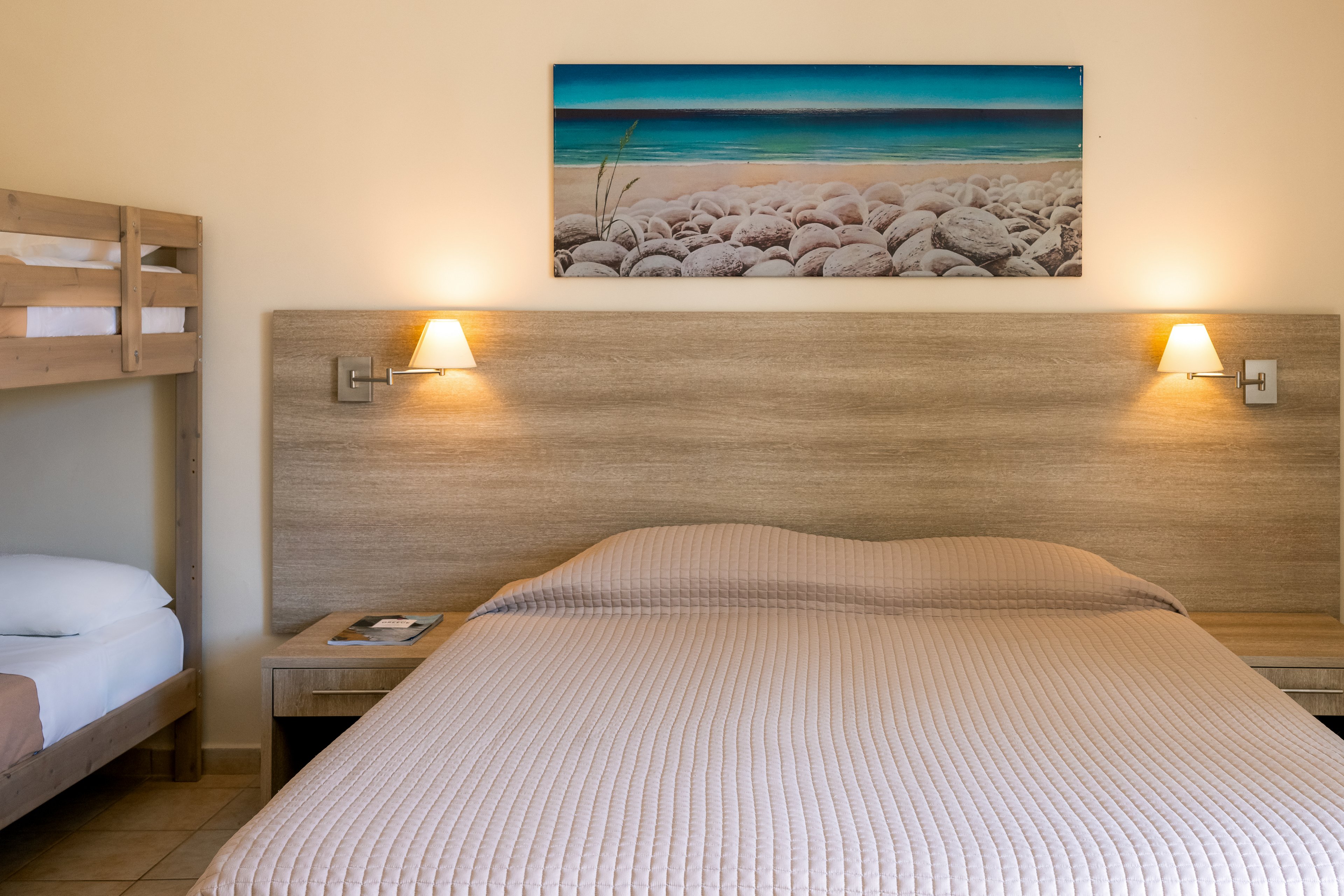 A well-lit hotel room featuring a double bed and bunk beds. The setting includes warm lighting and a serene beach-themed artwork above the bed. Neutral tones dominate the decor, creating a calm and inviting atmosphere.