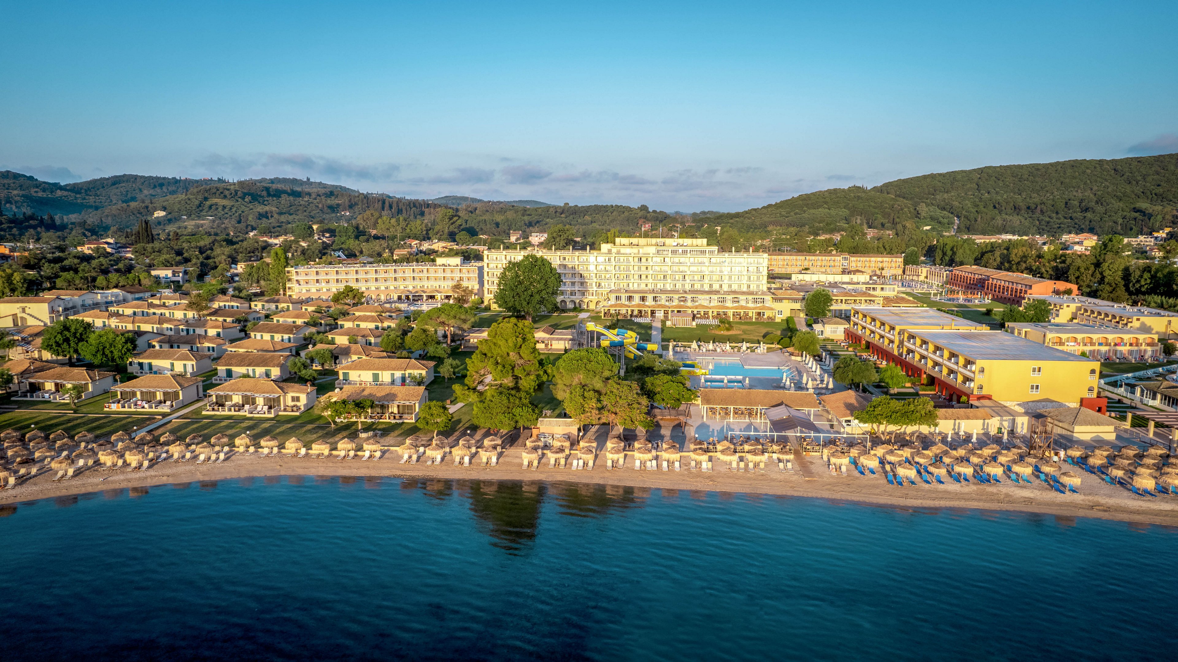 An expansive seaside resort is captured from above along a calm shoreline. Rows of sun loungers and umbrellas line the sandy beach beside the clear blue water. Behind the beach, multiple hotel buildings, pools, and sports courts are surrounded by greenery. Rolling hills and forested landscape form a scenic backdrop under a clear morning sky.