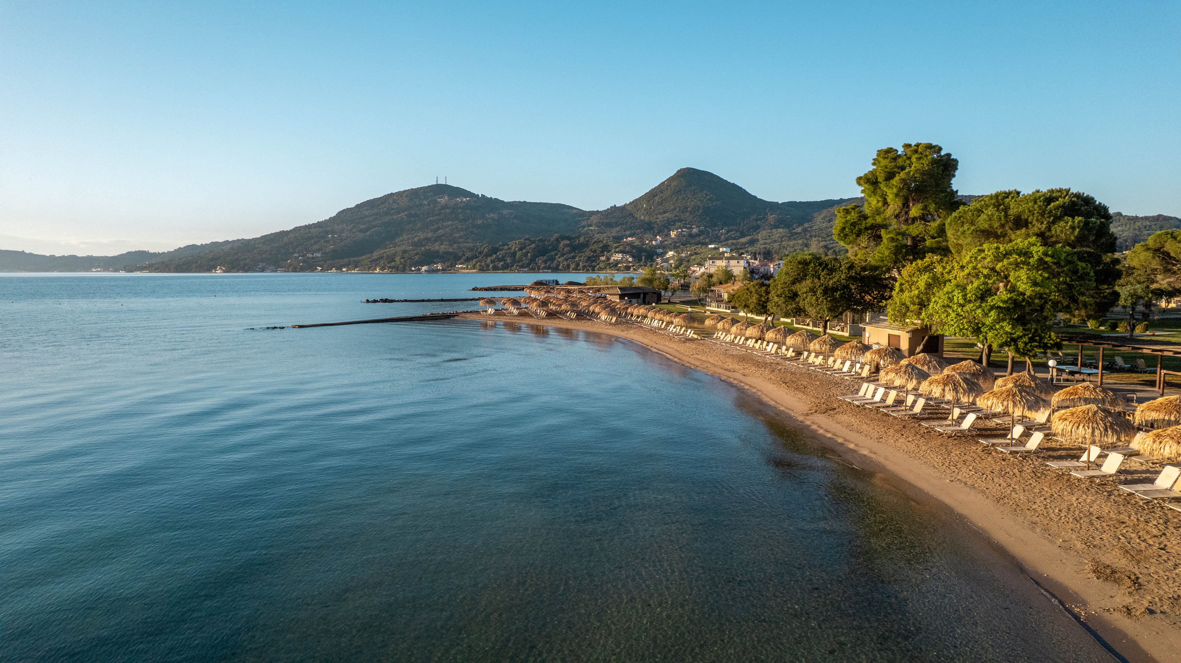 A tranquil sandy beach curves gently along a calm sea under clear blue skies. Neatly arranged sun loungers and straw parasols line the shoreline, suggesting a resort setting ready for visitors. Green hills and scattered buildings rise in the background, adding depth to the coastal landscape. The soft morning or late afternoon light creates a peaceful and inviting atmosphere.