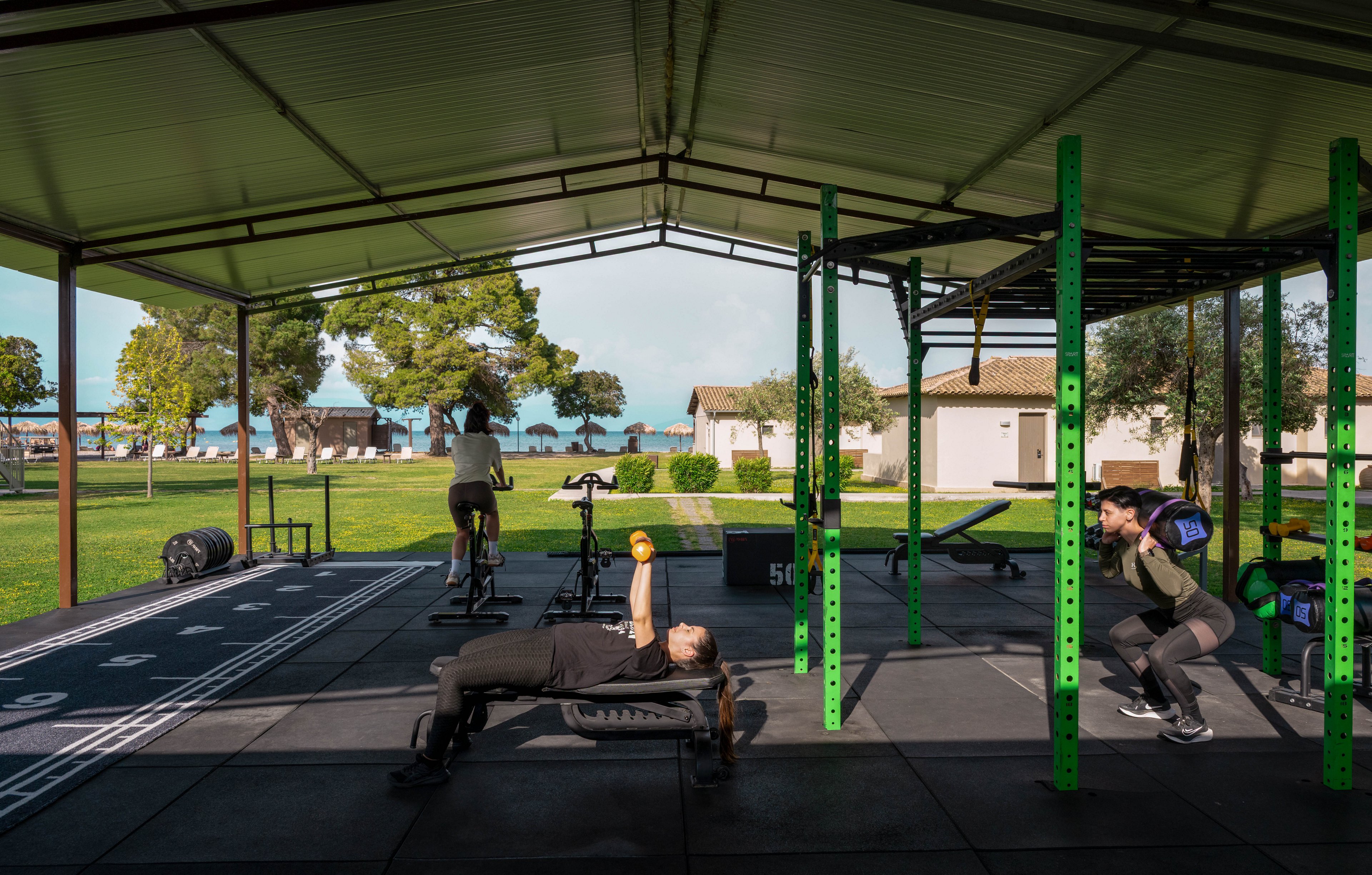 An outdoor covered gym features several people engaged in different exercises. One person performs a bench press on a step platform while another does squats with a barbell near a green metal rack. In the background, a person cycles on a stationary bike and another area of the gym shows additional equipment. The gym is set in a grassy park-like environment with trees, small buildings, and a view of water in the distance. Bright daylight and open sides of the structure create an airy, energetic workout atmosphere.