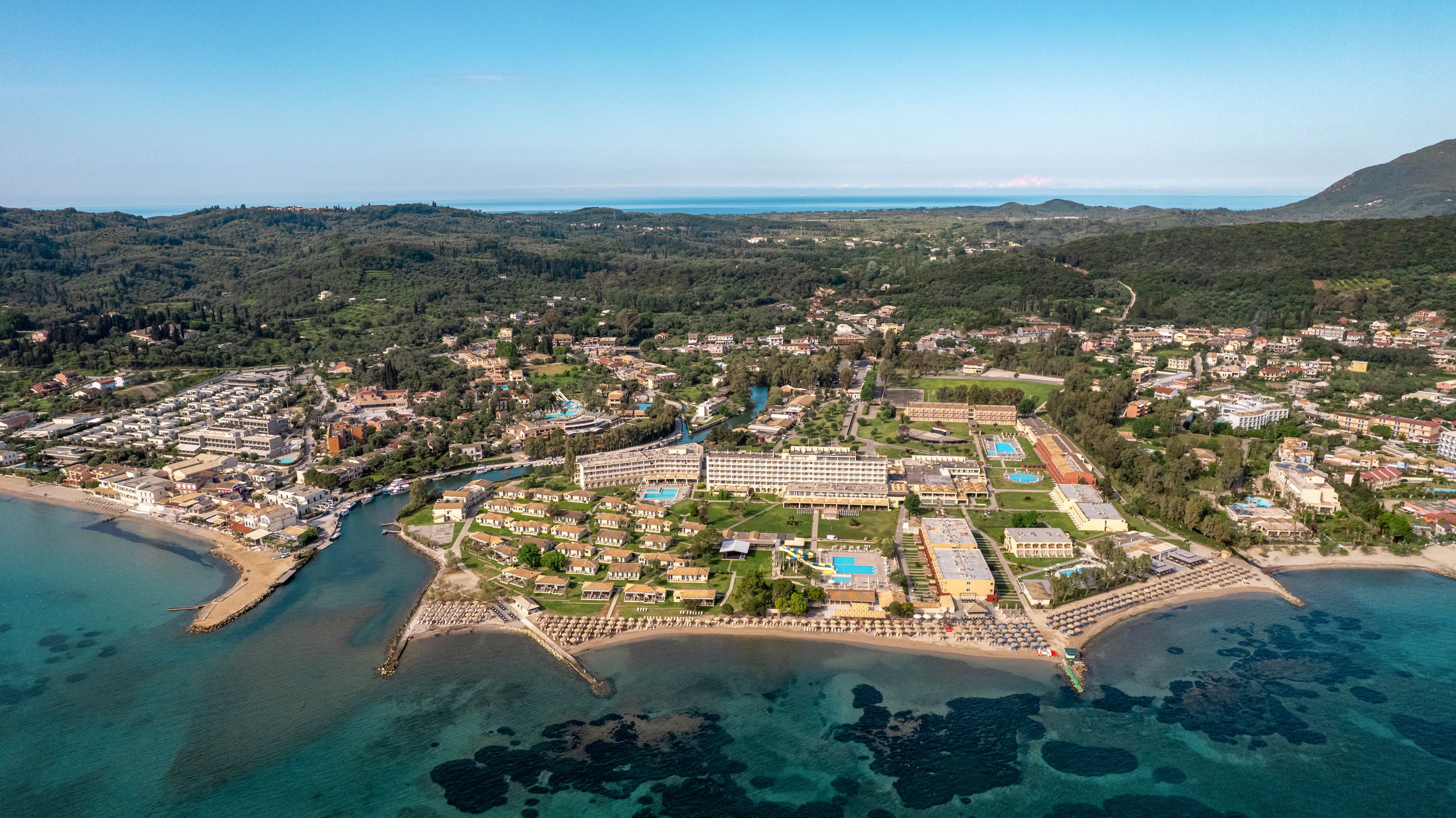 A large seaside resort complex stretches along a clear blue coastline, captured from an elevated aerial perspective. Multiple hotel buildings, swimming pools, and sports courts are arranged in neat rows near the shore. Behind the resort, a lush green landscape with scattered houses and hills extends toward the horizon. The calm sea in the foreground reveals shallow reefs and darker patches of underwater rock formations.