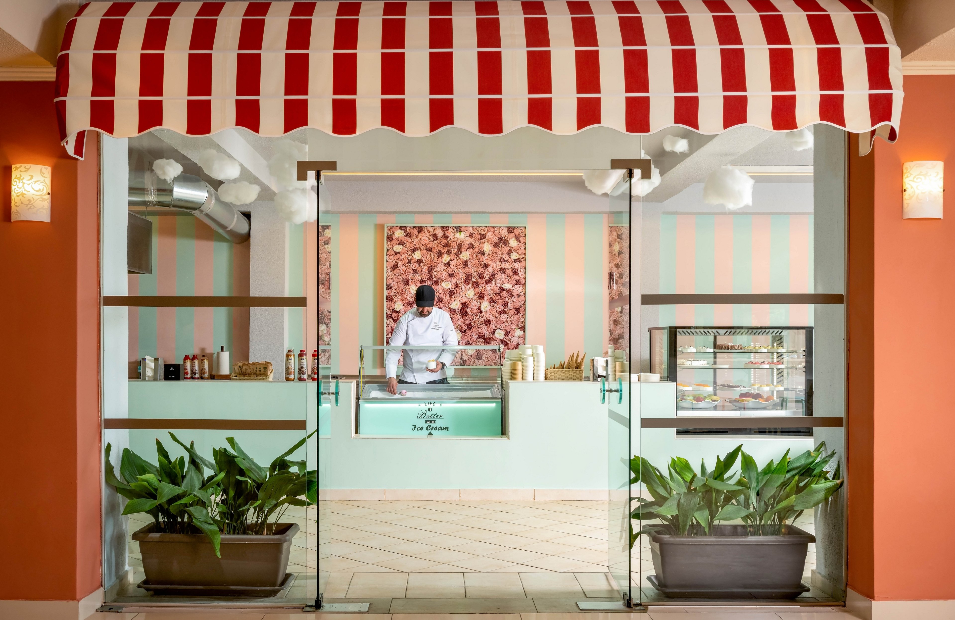 A small ice cream shop is viewed from the front through glass doors, framed by a red and white striped awning. Inside, a worker wearing a white chef coat and dark face mask prepares items behind a pastel green counter. The interior features mint green and peach vertical stripes on the walls and a floral artwork in the background. Display cases, bottles, and containers line the counter, while potted plants sit outside the entrance, adding greenery to the scene.