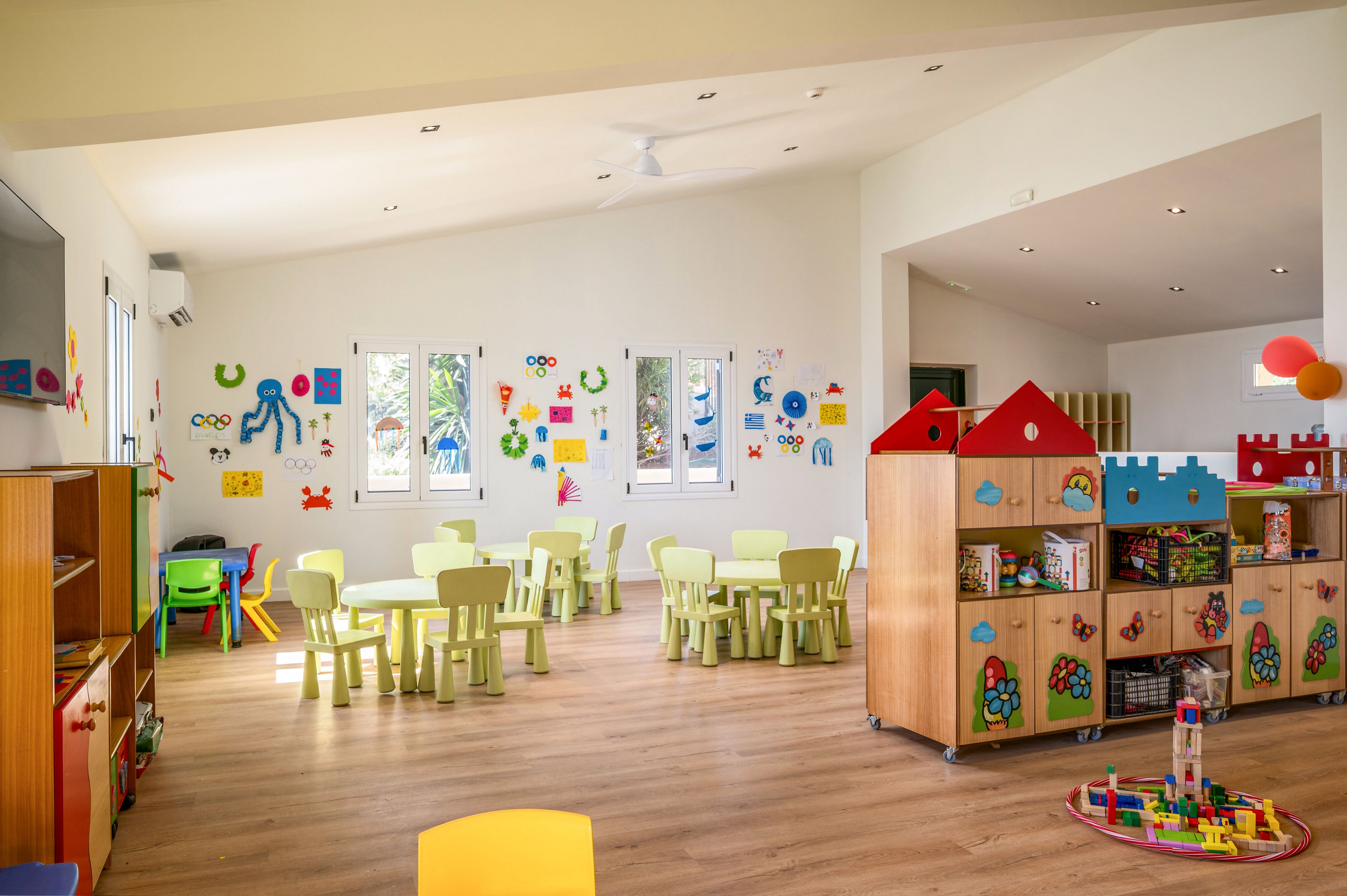 Spacious kindergarten classroom filled with small tables, chairs, and play areas. Wooden storage units hold various toys, blocks, and games, while a round play mat sits on the floor. The walls are decorated with colorful shapes, animals, and playful graphics around several windows. Natural light and soft colors create a cheerful, welcoming environment for young children.