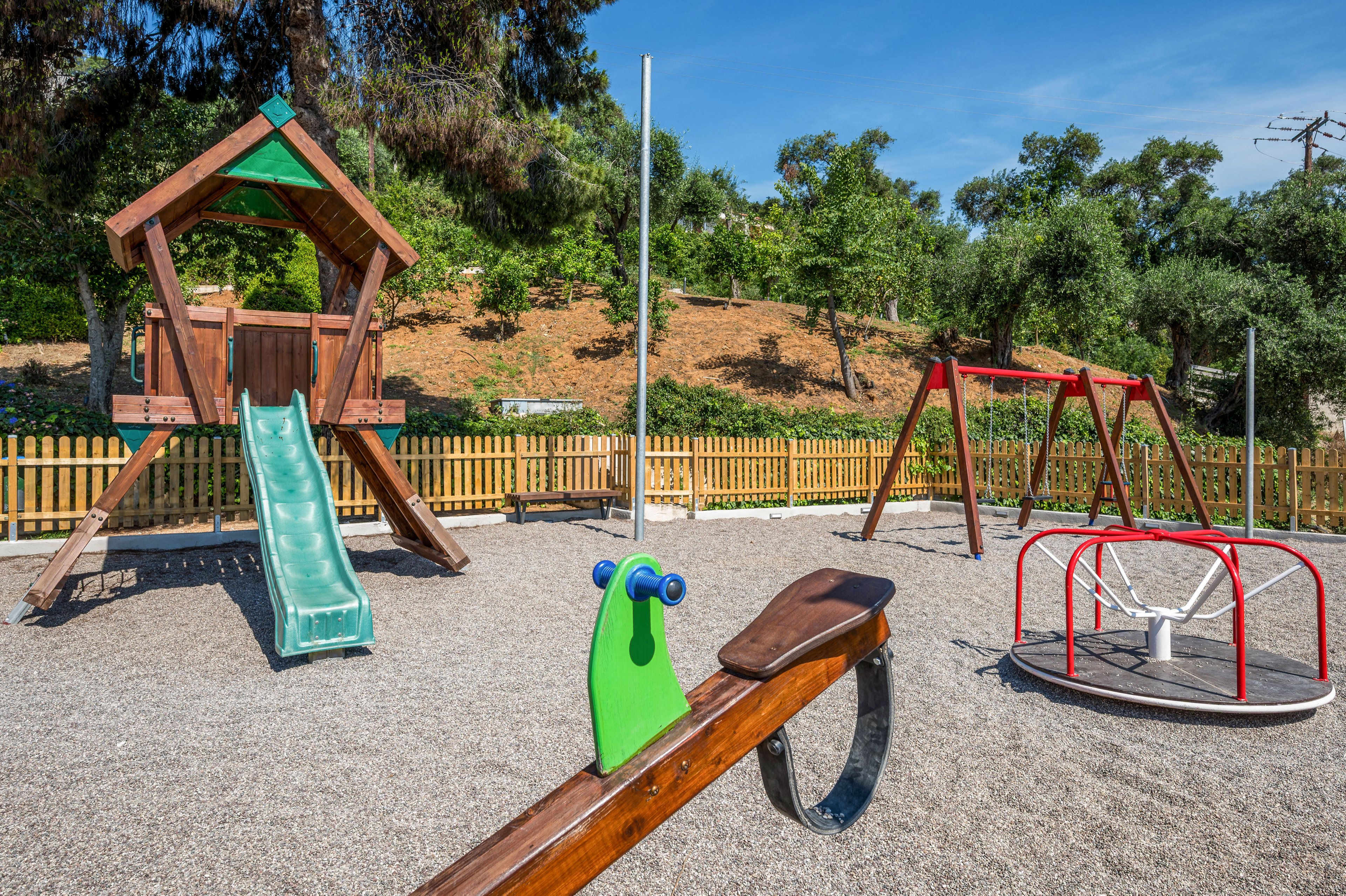 A colorful outdoor playground stands empty on a sunny day. The play area features a wooden playhouse with a green slide, a seesaw, a swing set, and a round merry-go-round on a gravel surface. A wooden fence and lush green trees surround the playground, creating a safe and natural environment. Bright daylight and clear skies give the scene a cheerful and inviting atmosphere.