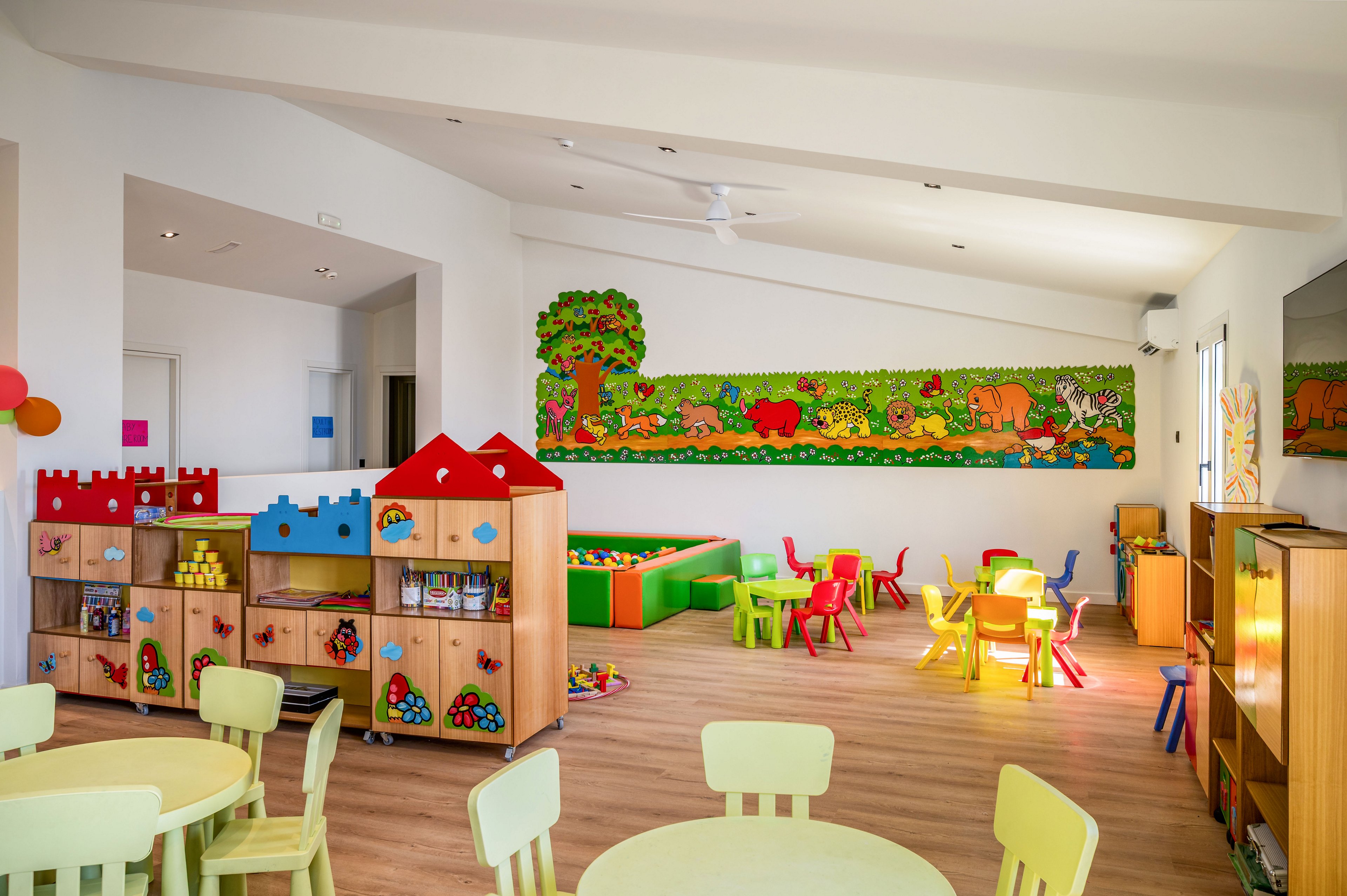 Bright, spacious kindergarten classroom featuring small tables and chairs arranged for group activities. A central play area with soft blocks and colorful furniture is surrounded by wooden storage units filled with toys and books. The walls are decorated with a long, vibrant mural of cartoon animals and trees, creating a cheerful atmosphere. Natural and artificial light enhance the playful colors and clean, organized layout of the room.