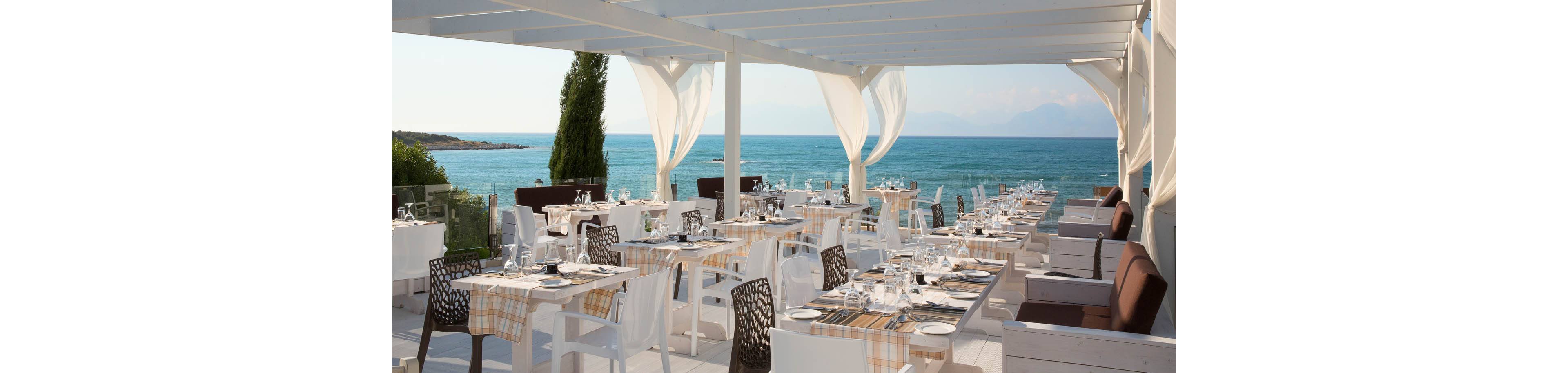 Open-air restaurant tables are neatly arranged on a terrace overlooking a calm blue sea. White pergola beams and flowing curtains frame the coastal view, creating a bright and airy dining space. Tables are set with glassware, cutlery, and checkered tablecloths, ready for guests. Modern white and dark woven chairs add contrast to the serene, sunlit setting.