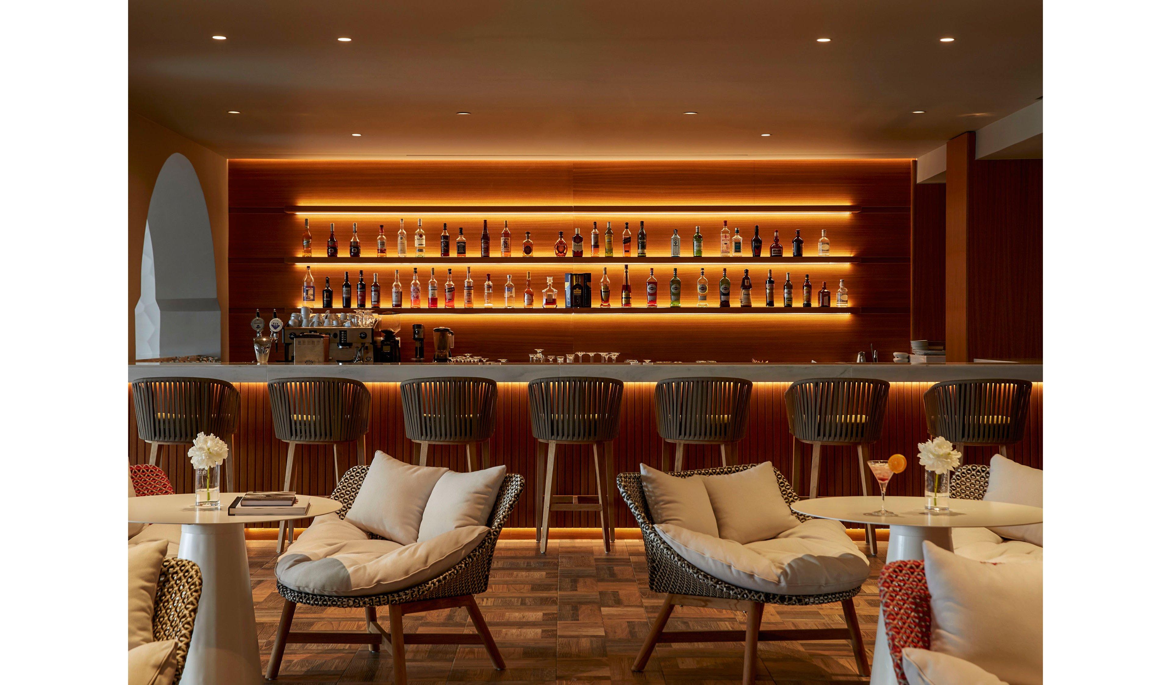 A stylish bar interior features a long counter with modern high stools and softly glowing backlit shelves displaying assorted bottles. In the foreground, cushioned lounge chairs and small tables with white flowers create a relaxed seating area. The warm, golden lighting contrasts with neutral upholstery and wooden textures, giving the space an inviting, upscale atmosphere. The overall scene suggests a sophisticated hotel or resort bar designed for evening socializing and casual drinks.