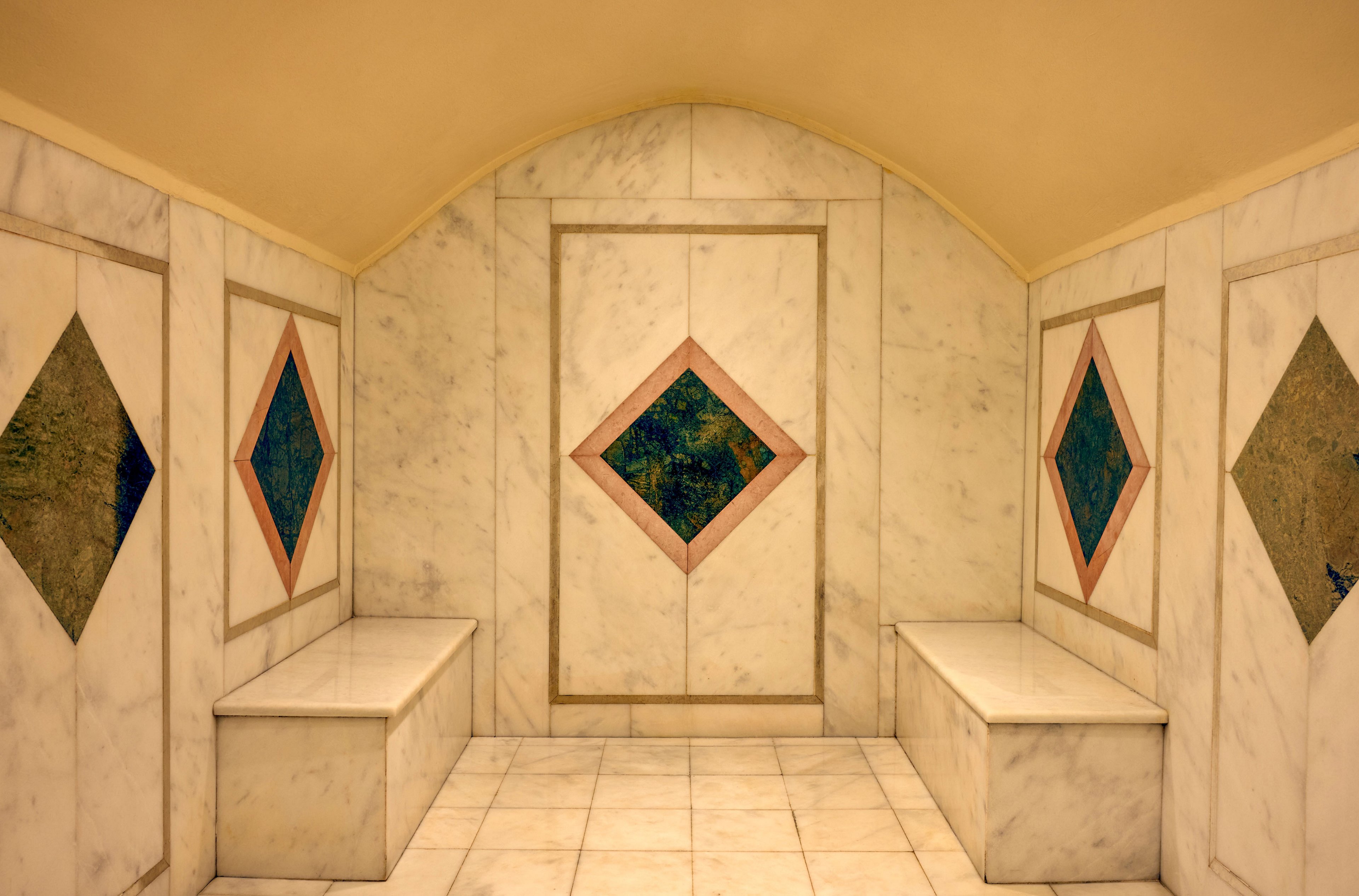 Symmetrical marble room features two bench seats and a central wall panel. Diamond-shaped insets with colorful stone or tile accents decorate the side and center walls. The arched ceiling and light-toned marble surfaces create a clean, refined atmosphere. The tiled floor and geometric design suggest a spa, bath, or decorative interior space.