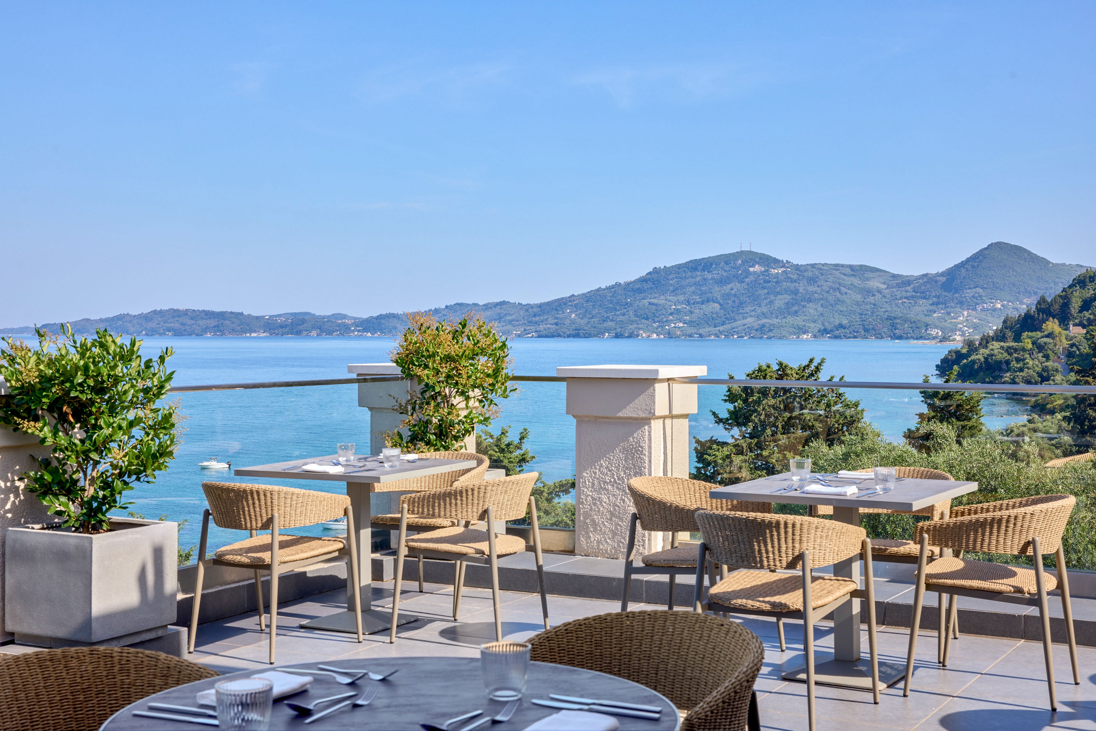 An elegant outdoor restaurant terrace overlooks a calm blue sea and distant green hills. Several tables and wicker chairs are neatly arranged, set with plates, glasses, and cutlery for guests. Potted plants and a glass railing frame the view, adding greenery to the modern setting. The bright daylight and clear sky create a relaxed, vacation-like atmosphere.