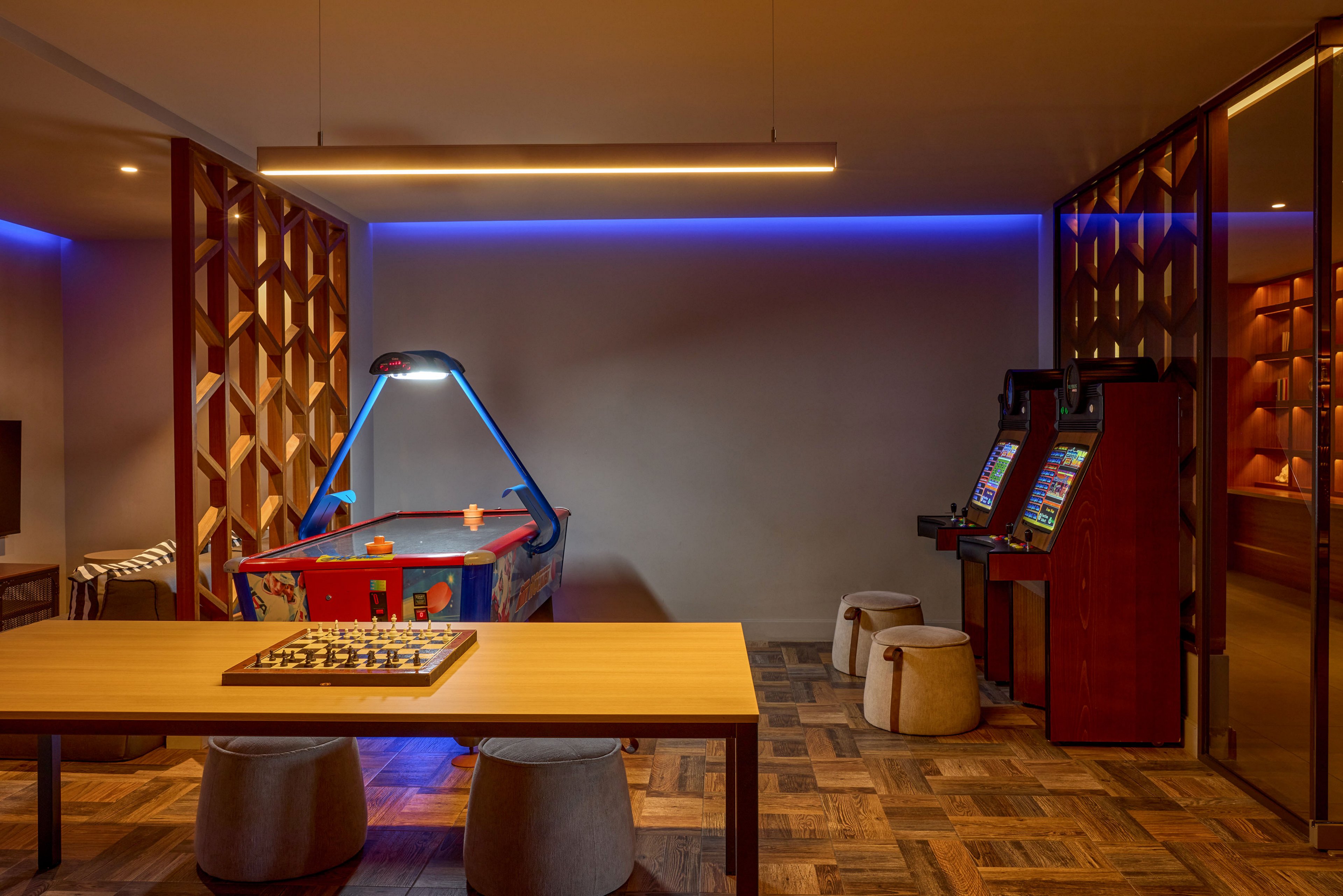 A contemporary game room features an air hockey table illuminated by a blue overhead light. In the foreground, a wooden table holds a chessboard, surrounded by low cushioned stools on a patterned wood floor. To the right, two upright arcade machines stand against the wall, adding a retro gaming touch. Warm ambient lighting and decorative wooden partitions create a cozy, inviting atmosphere.