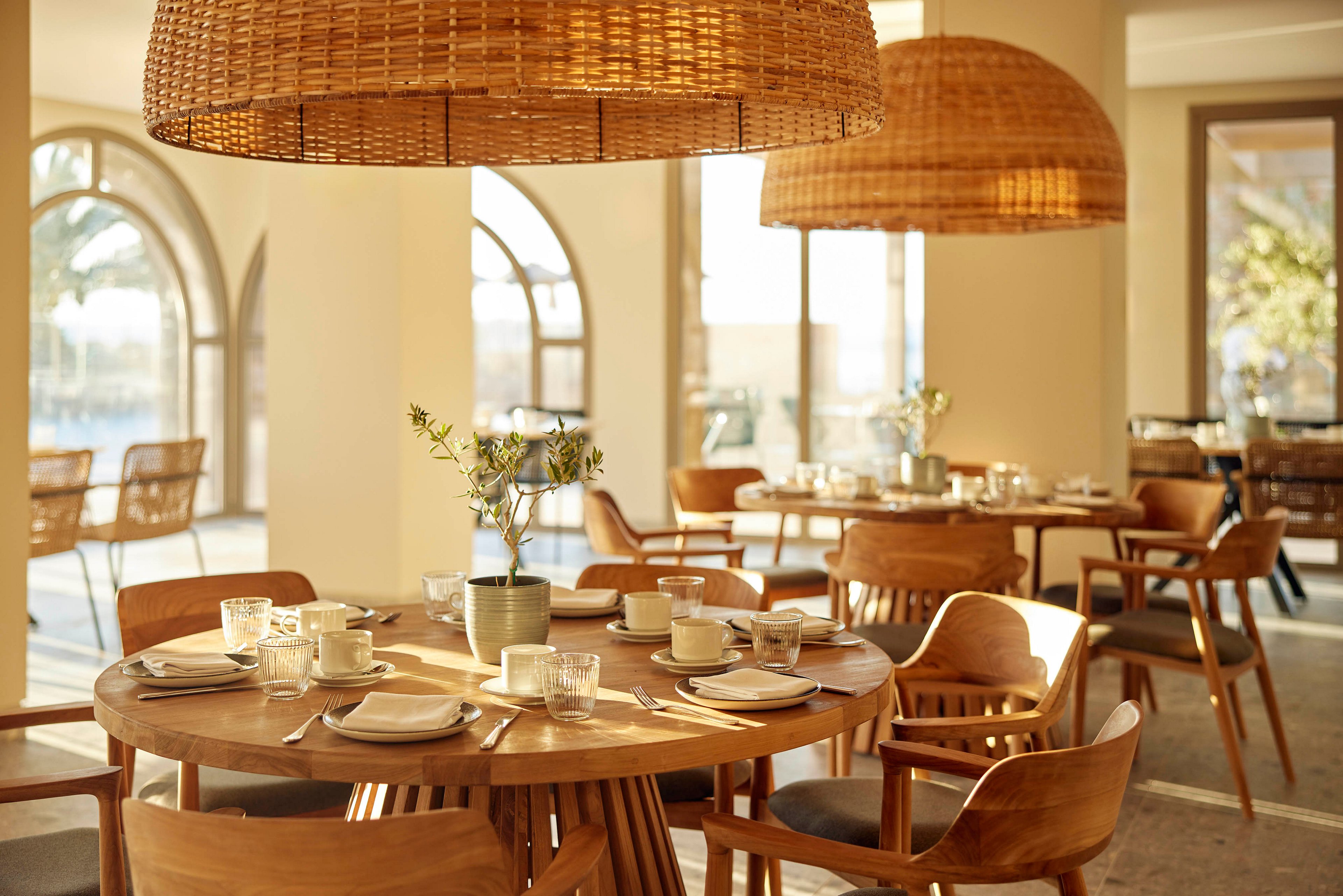 A bright, contemporary restaurant interior features a round wooden table set with plates, cups, glasses, and cutlery. Large woven pendant lights hang overhead, casting warm light across the room. Wooden chairs and additional tables fill the space, framed by tall arched windows that let in abundant natural sunlight. A small potted plant centerpiece adds a touch of greenery to the calm, inviting atmosphere.