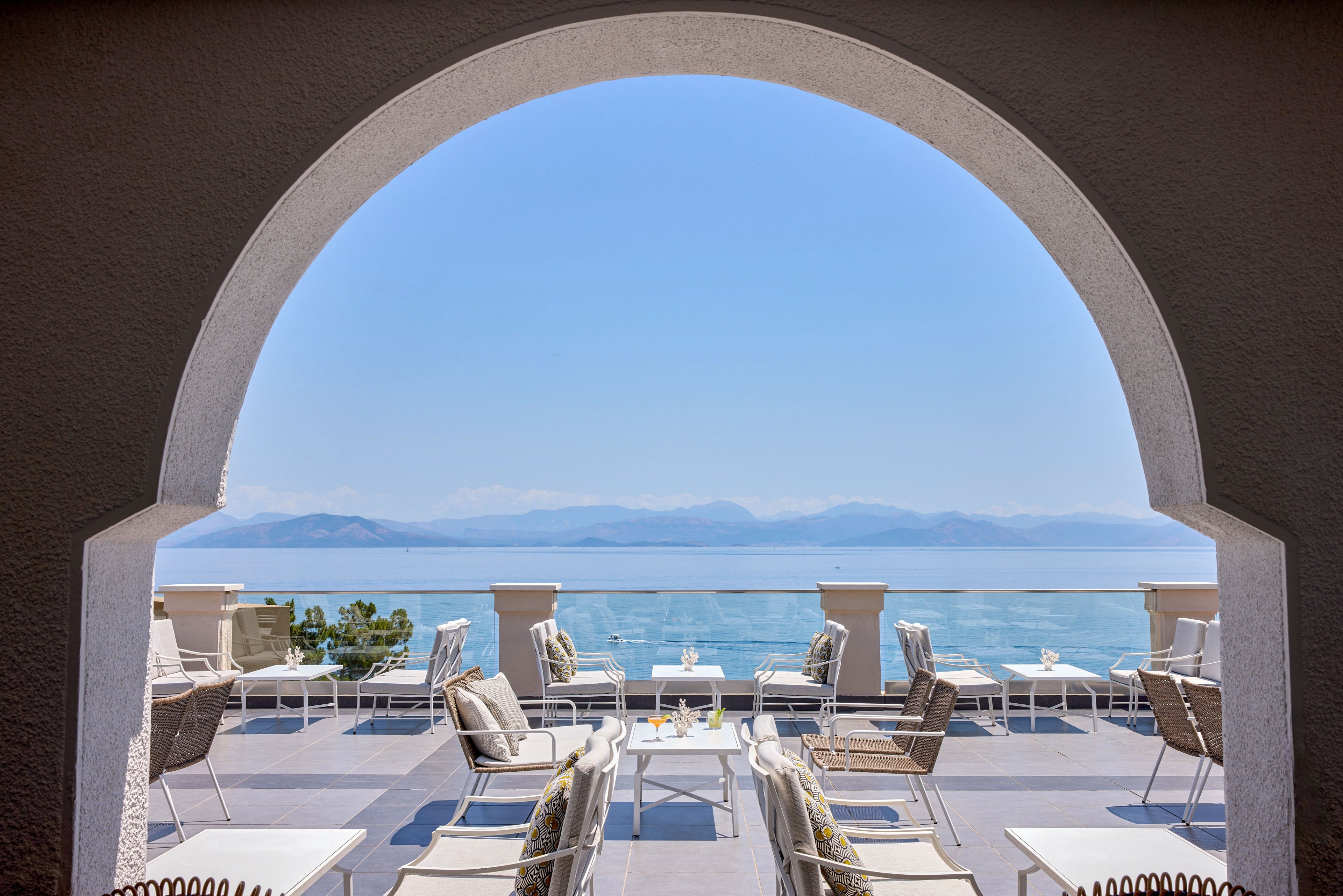 An elegant outdoor lounge terrace overlooks a calm, expansive sea framed by a large arched opening. White lounge chairs and cushioned seating are arranged around low tables, some holding drinks and glassware. The scene suggests a resort or hotel setting with a serene, relaxing atmosphere. Distant mountains and a clear blue sky enhance the coastal, vacation feel.