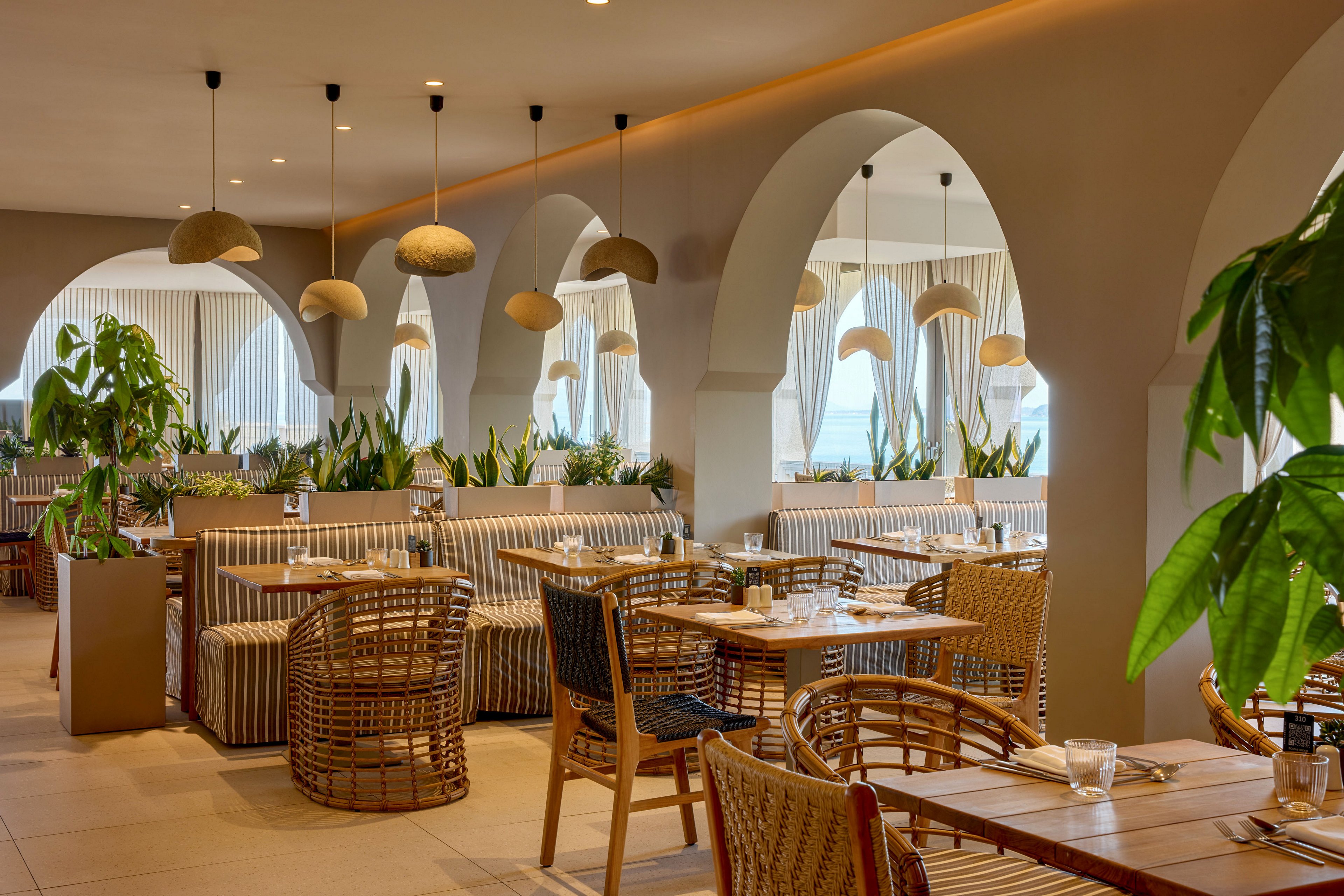 Contemporary restaurant dining area featuring arched walls and warm lighting. Wooden tables and woven chairs are arranged neatly alongside striped upholstered banquettes. Green potted plants add natural accents throughout the space, complementing the neutral color palette. Large windows with sheer curtains suggest a bright, airy atmosphere suitable for relaxed dining.