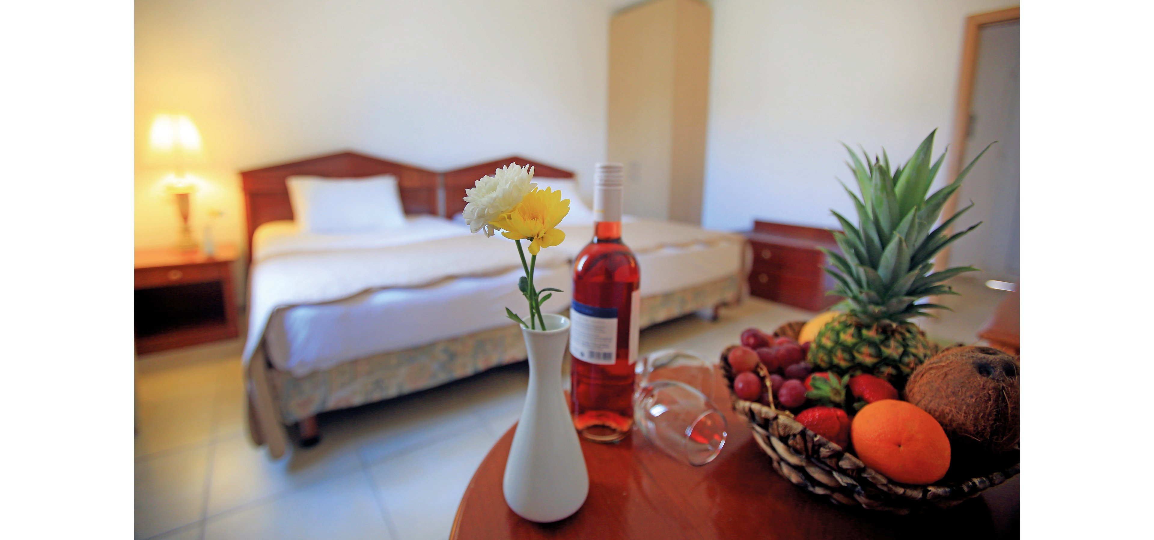 A well-lit hotel room featuring a neatly made bed and a wooden table in the foreground. The table displays a bottle of rose wine, an empty wine glass, a vase with fresh flowers, and a basket of assorted fruits including pineapple, coconut, and oranges. The warm lighting and simple decor create a welcoming and relaxing atmosphere.