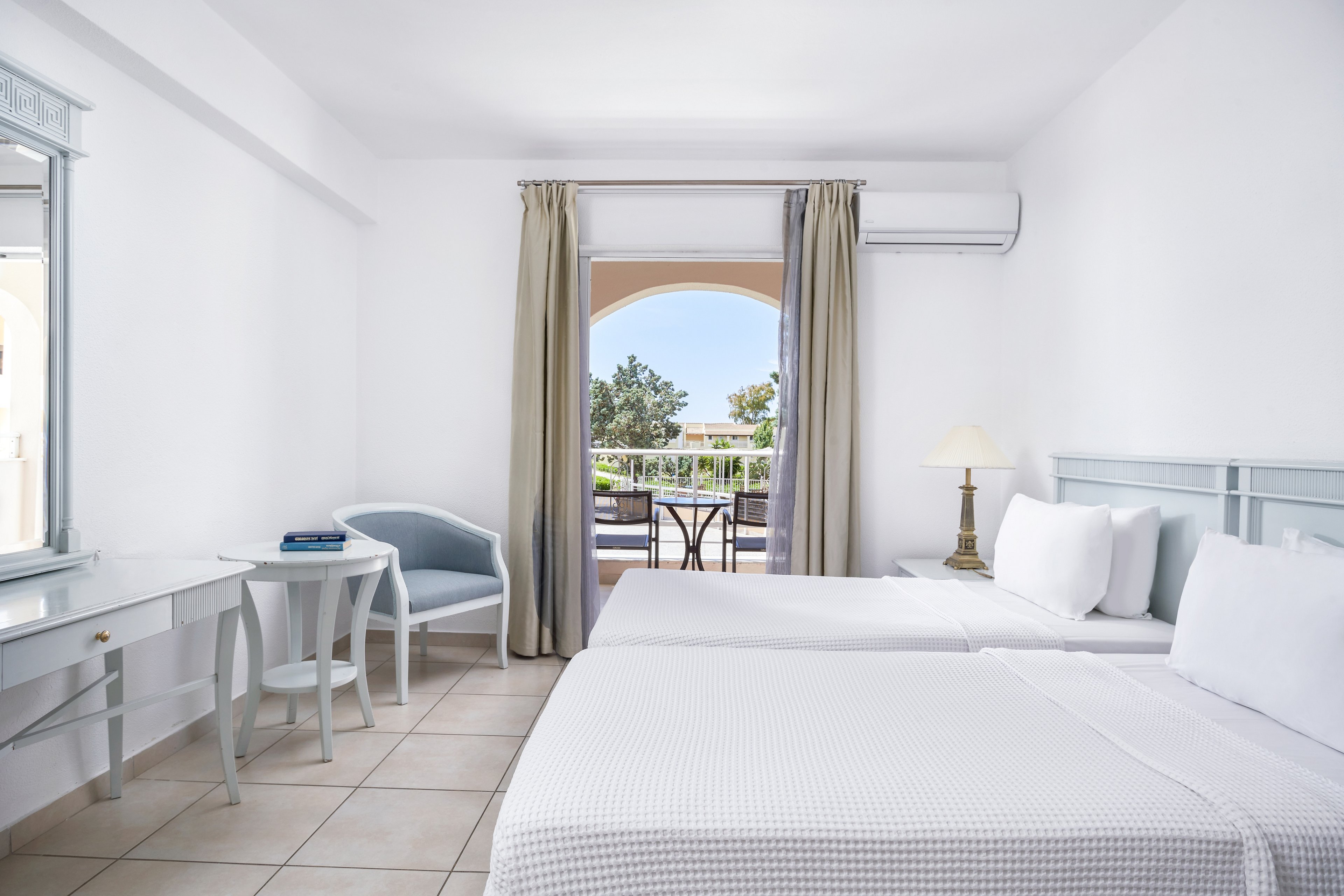 A spacious and bright hotel room featuring two neatly made twin beds, a desk with a mirror, and a small seating area. The room opens up to a balcony with a view of greenery and clear skies. The decor is minimalist with white walls, light furniture, and soft natural lighting.