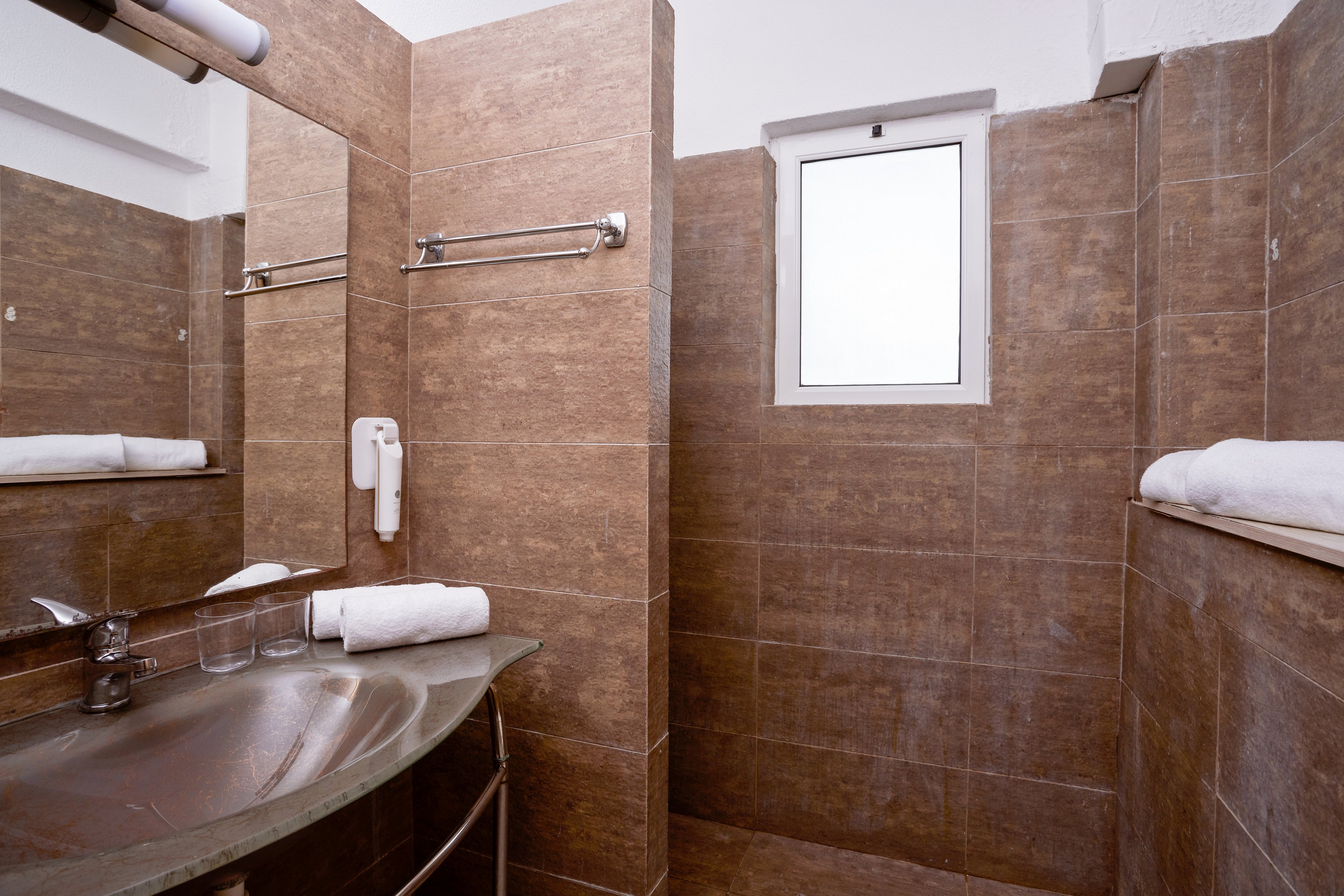 A contemporary bathroom featuring brown tiled walls and a sleek glass sink. The setting includes a towel rack, folded white towels, and a mounted hairdryer. A small window provides natural light, enhancing the clean and minimalist design.