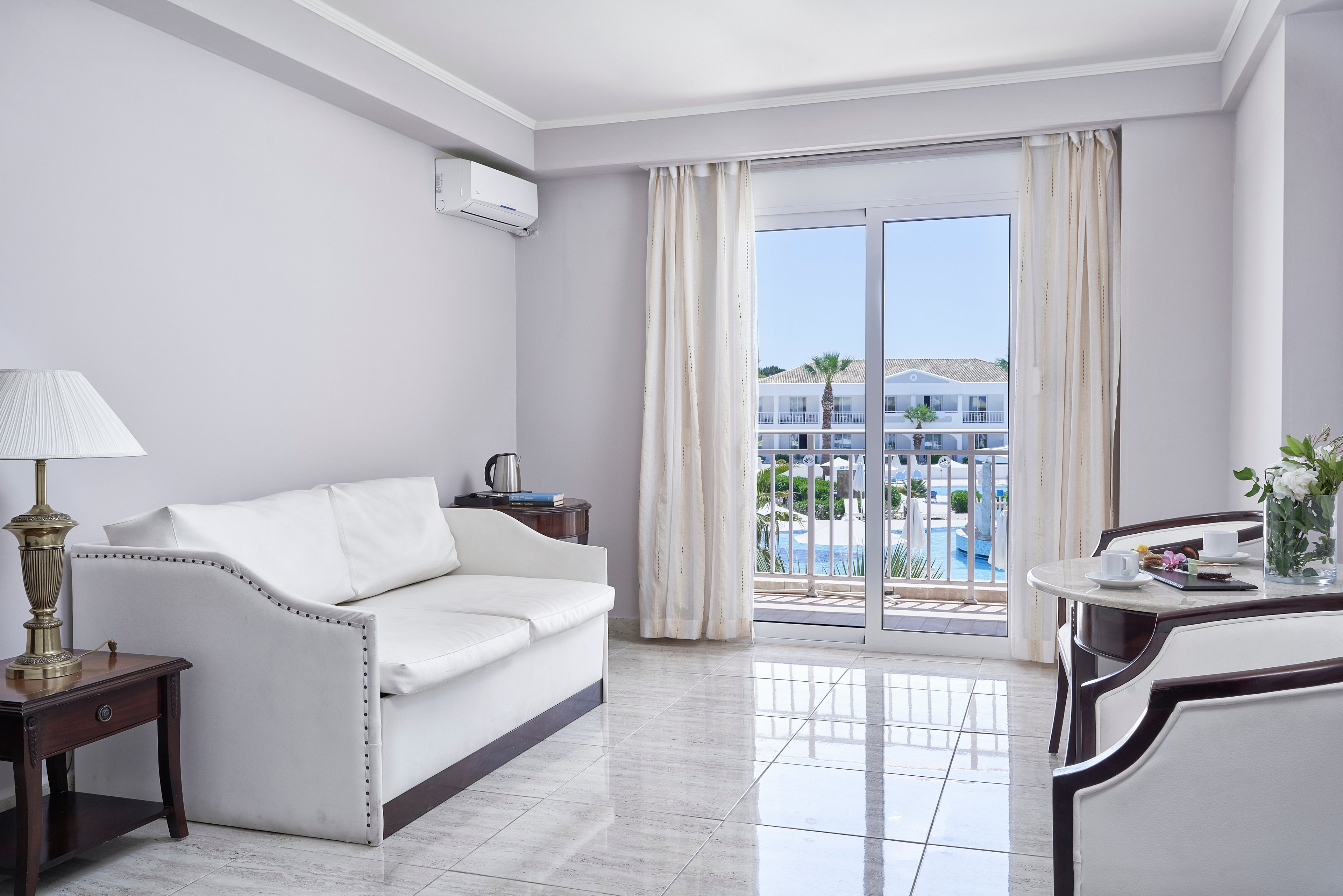 A modern hotel room featuring a white sofa, wooden furniture, and a small table setup. The room is well-lit with natural light streaming through large glass doors that open to a balcony overlooking a pool and resort area. Neutral tones dominate the decor, creating a calm and inviting atmosphere.