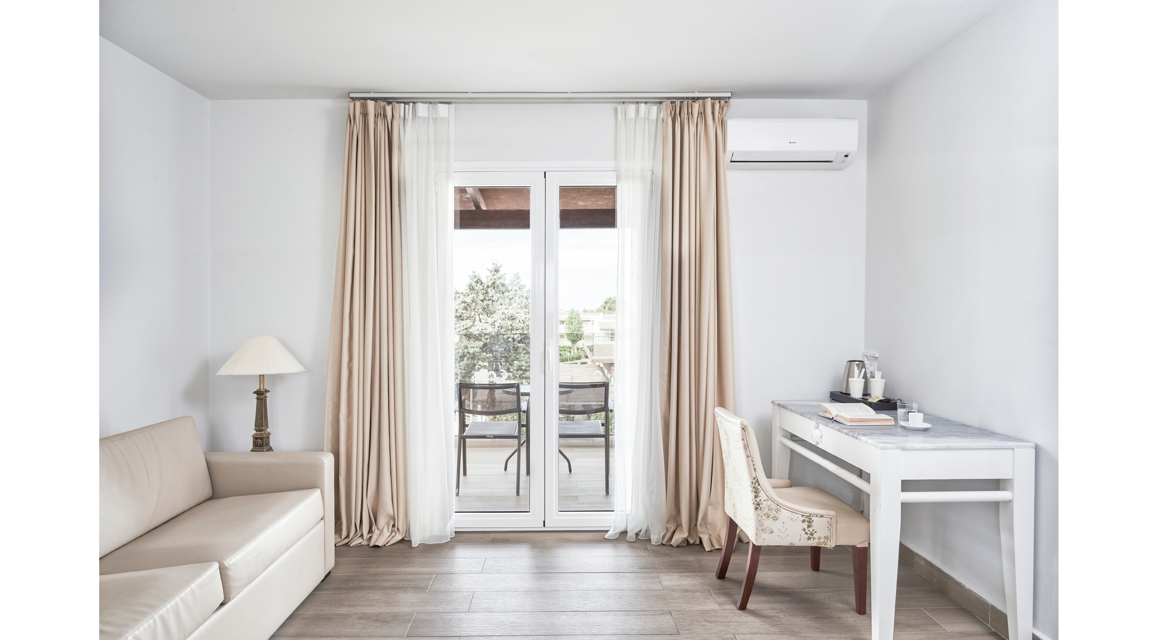 A minimalist and bright room featuring a beige sofa, a white desk with a chair, and a lamp. The setting includes large glass doors leading to a balcony with outdoor seating and greenery visible in the background. Neutral tones dominate the palette, complemented by soft curtains and wooden flooring.