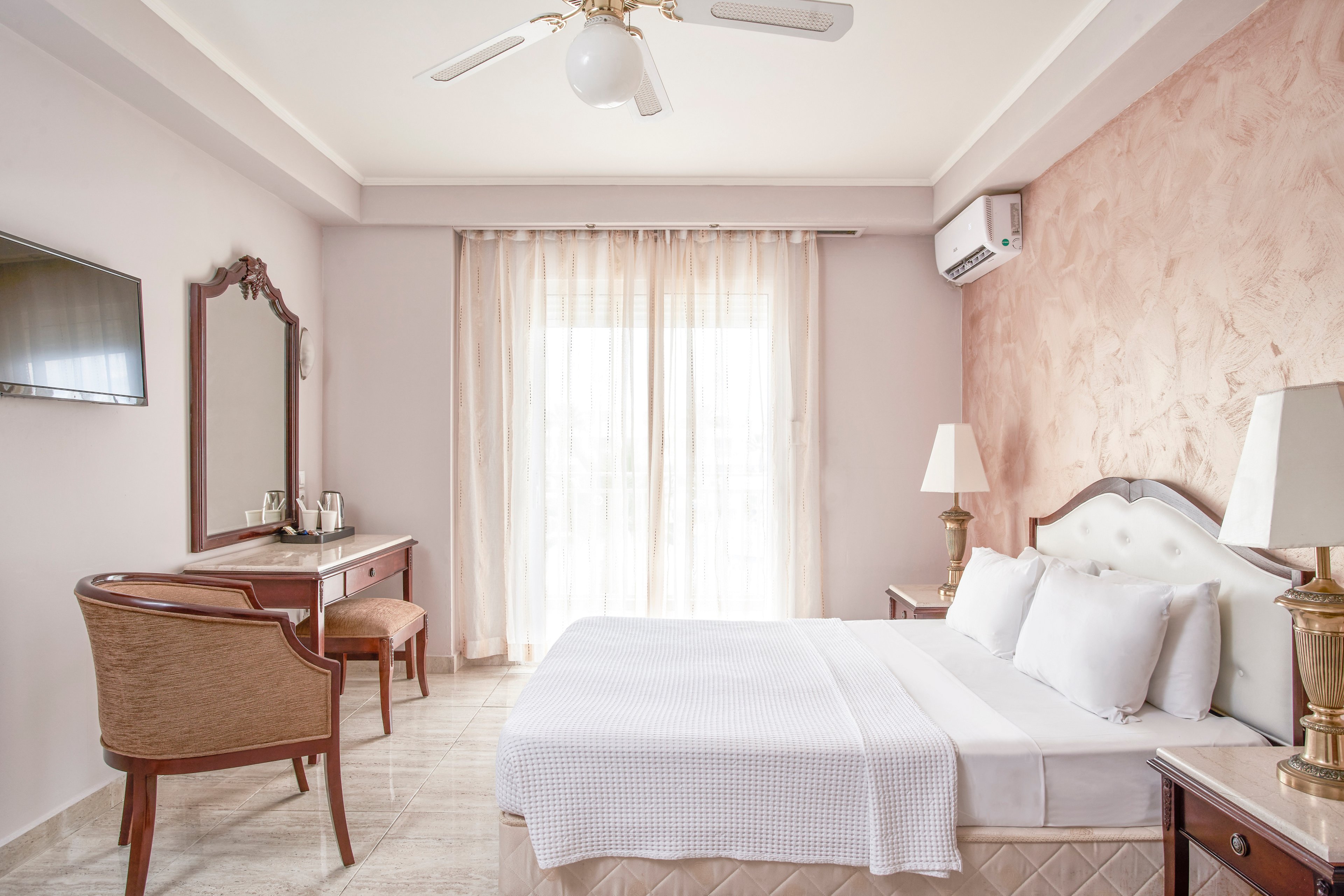 A well-lit hotel room featuring a double bed with white linens, a wooden desk and chair, and a wall-mounted TV. The room has a soft pink accent wall and large windows with sheer curtains allowing natural light to fill the space. An air conditioning unit is visible on the wall, ensuring comfort for guests.