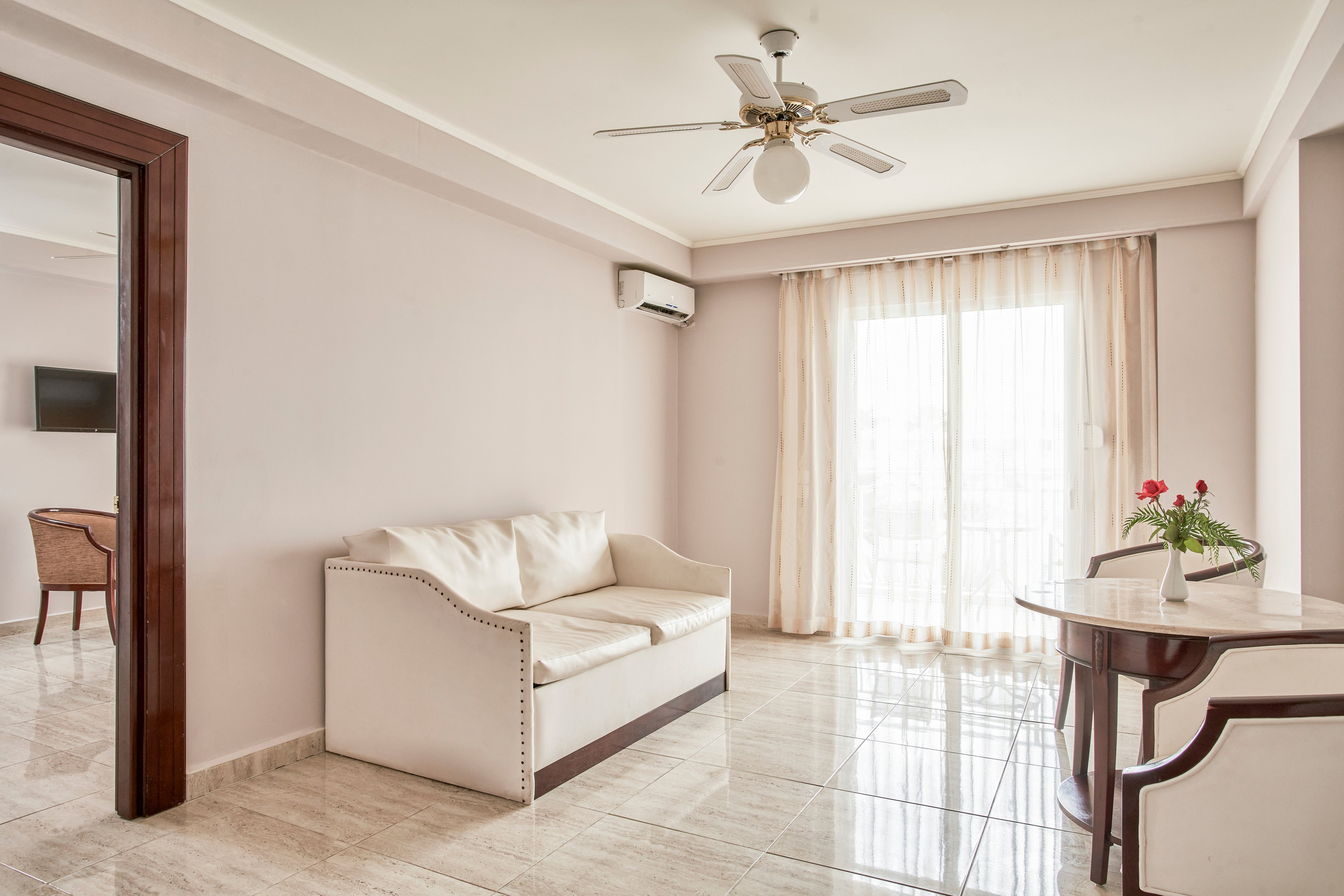 A spacious living room featuring a white sofa and a small table with a floral centerpiece. The room is well-lit with natural light streaming through sheer curtains covering a large glass door. A ceiling fan and air conditioning unit are visible, adding comfort to the minimalist decor. Neutral tones dominate the palette, creating a calm and inviting atmosphere.
