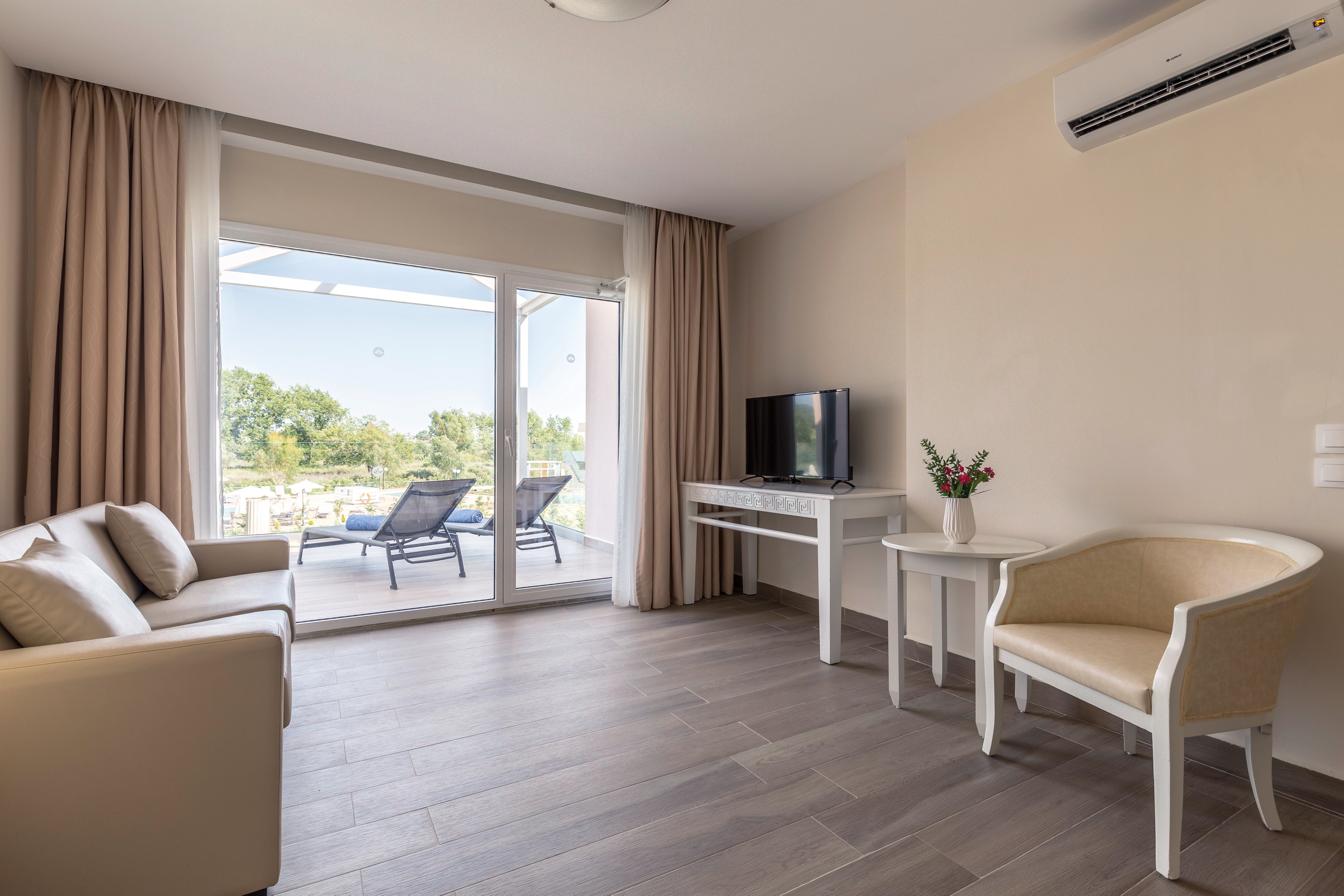 A bright and modern hotel room featuring a beige sofa, armchair, and a small desk with a television. The room opens to a patio with lounge chairs and greenery visible through large glass doors. Neutral tones dominate the decor, complemented by wooden flooring and soft curtains.