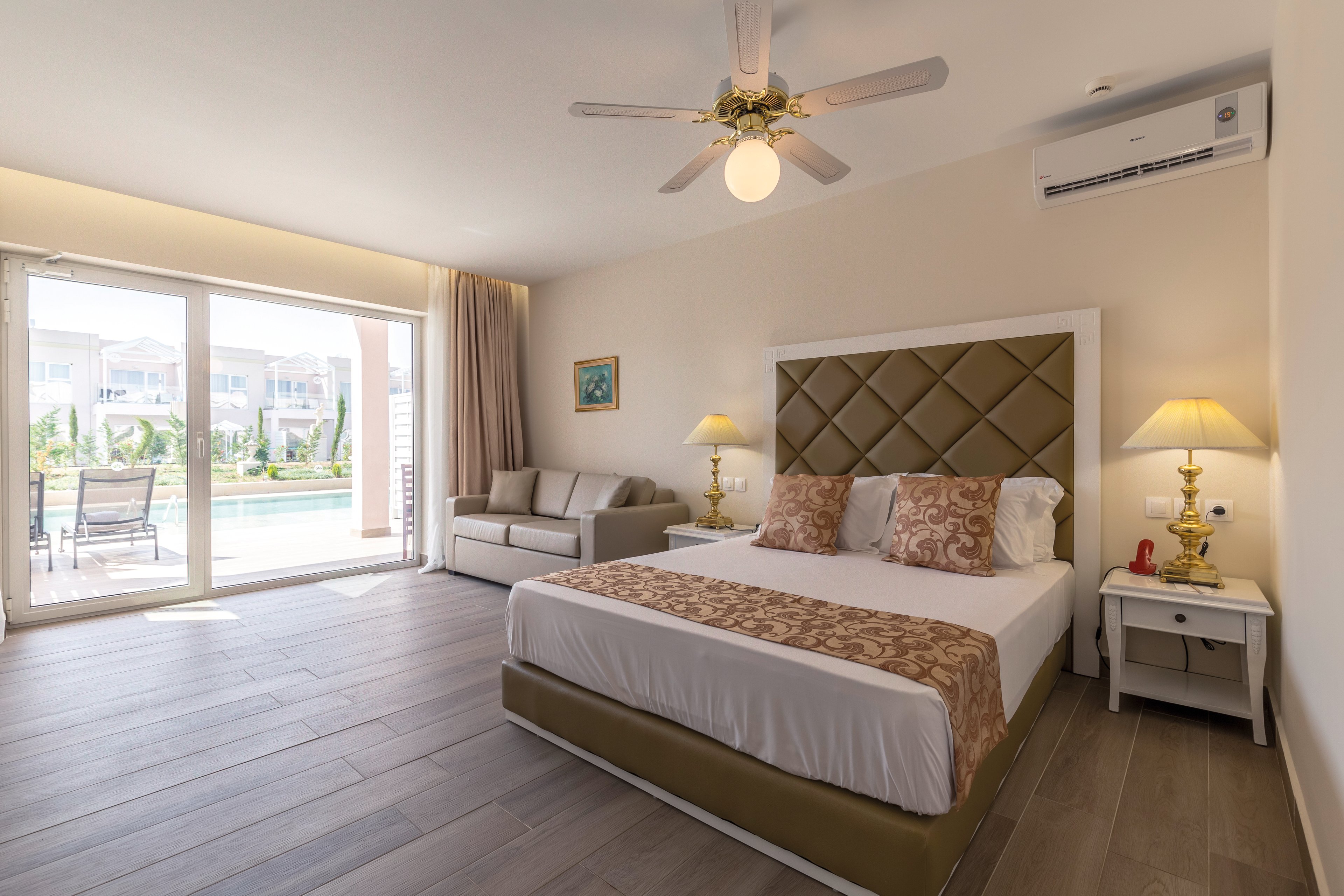 A spacious and well-lit hotel room featuring a large bed with a padded headboard and decorative pillows. The room includes a sofa, bedside tables with lamps, and a ceiling fan. A sliding glass door opens to a view of a pool and outdoor seating area. The air conditioning unit is visible on the wall.