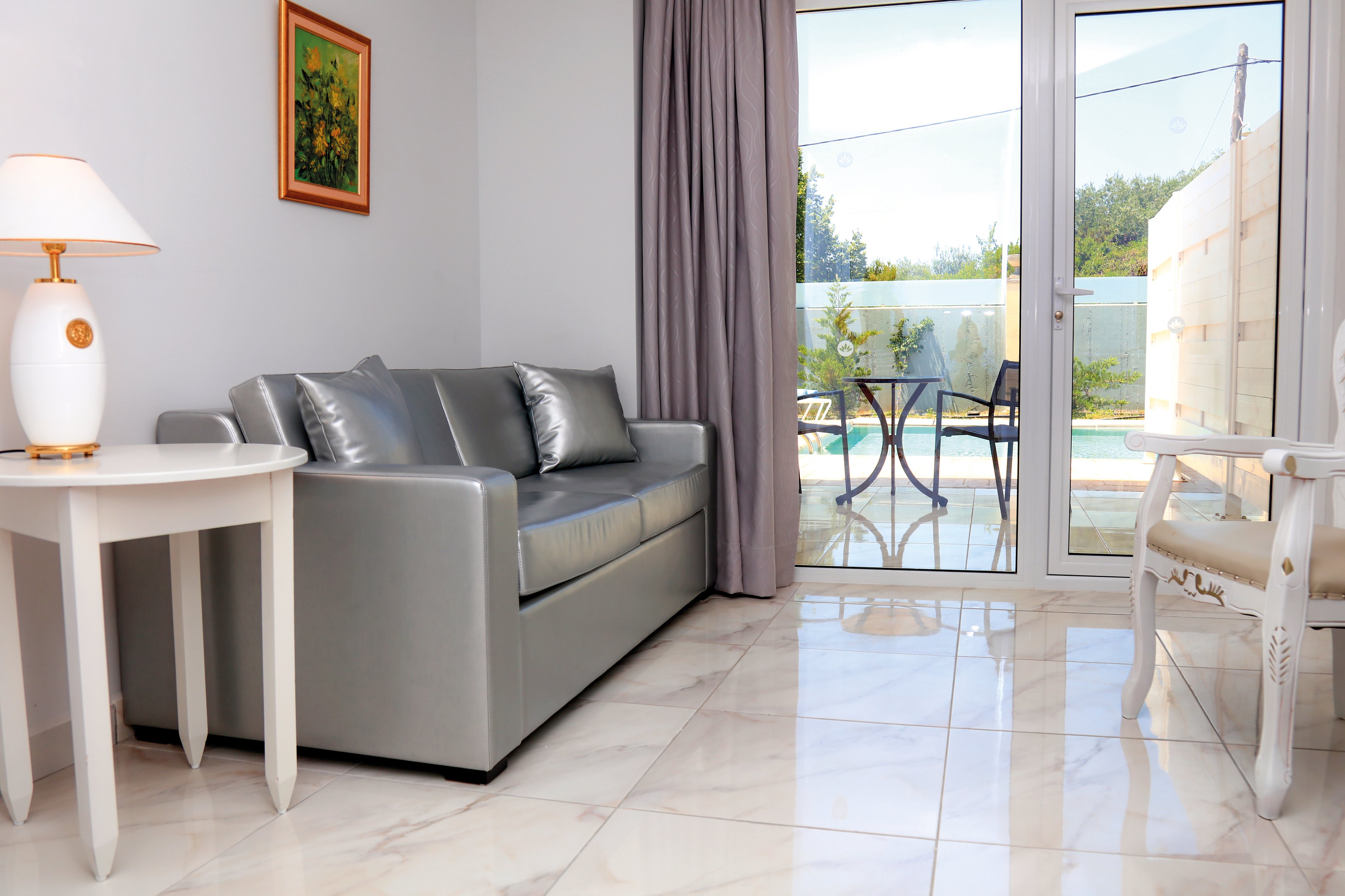 A contemporary living room featuring a sleek gray sofa and white furniture. The space is bright with natural light streaming through glass doors that open to a poolside patio. The setting includes a serene outdoor view with greenery and a small table and chairs visible outside.