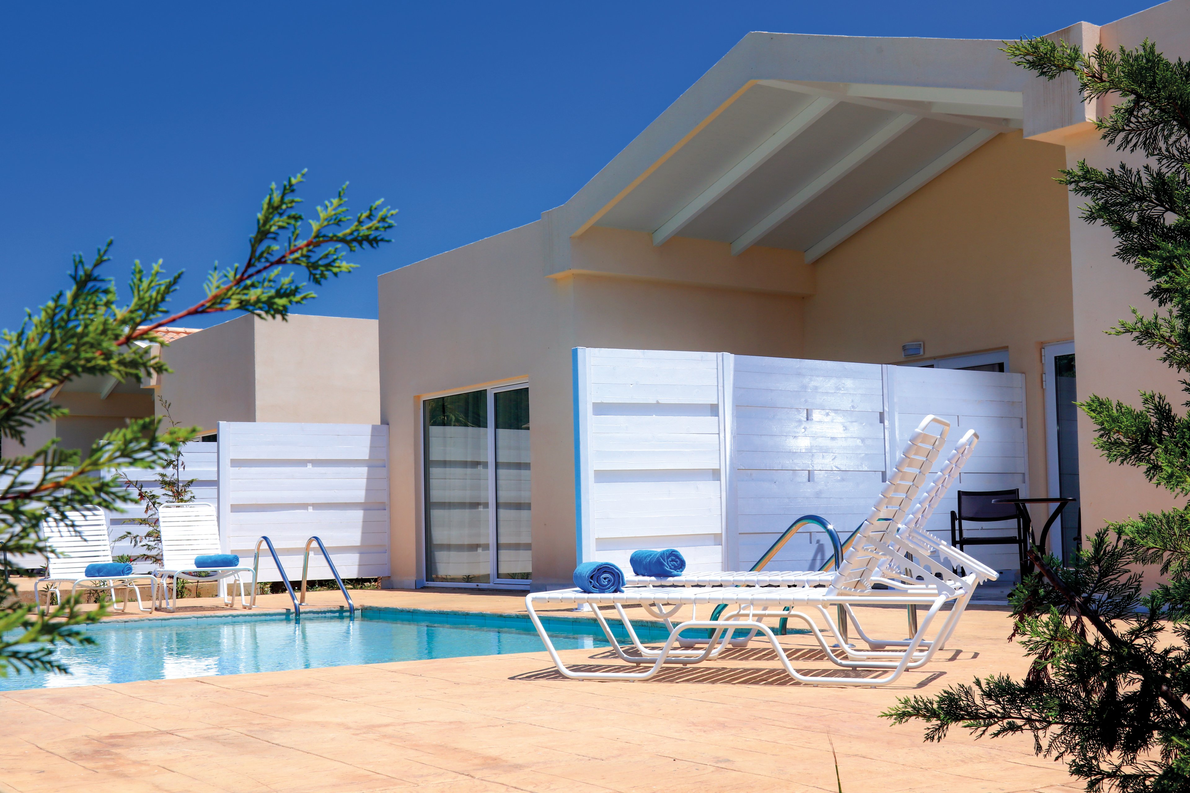 A contemporary villa featuring a private pool surrounded by lounge chairs and towels. The setting is outdoors under a clear blue sky, with minimalistic white architecture and greenery framing the scene. The area exudes relaxation and leisure, ideal for a sunny getaway.