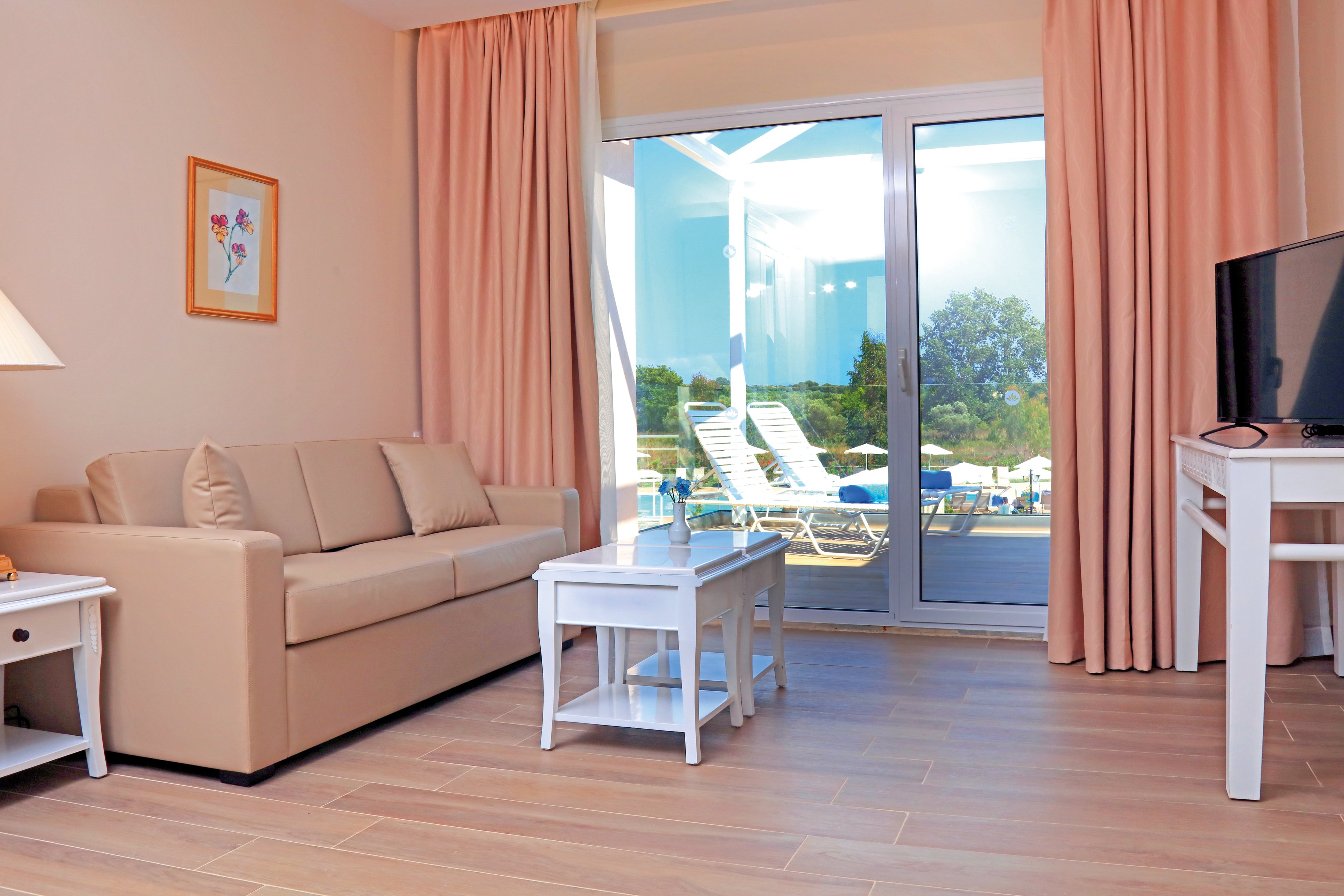 A bright and cozy living room featuring a beige sofa, white furniture, and soft pink curtains. The room opens to a balcony with lounge chairs and a scenic outdoor view of trees and blue skies. The setting is serene and inviting, ideal for relaxation or vacation stays.
