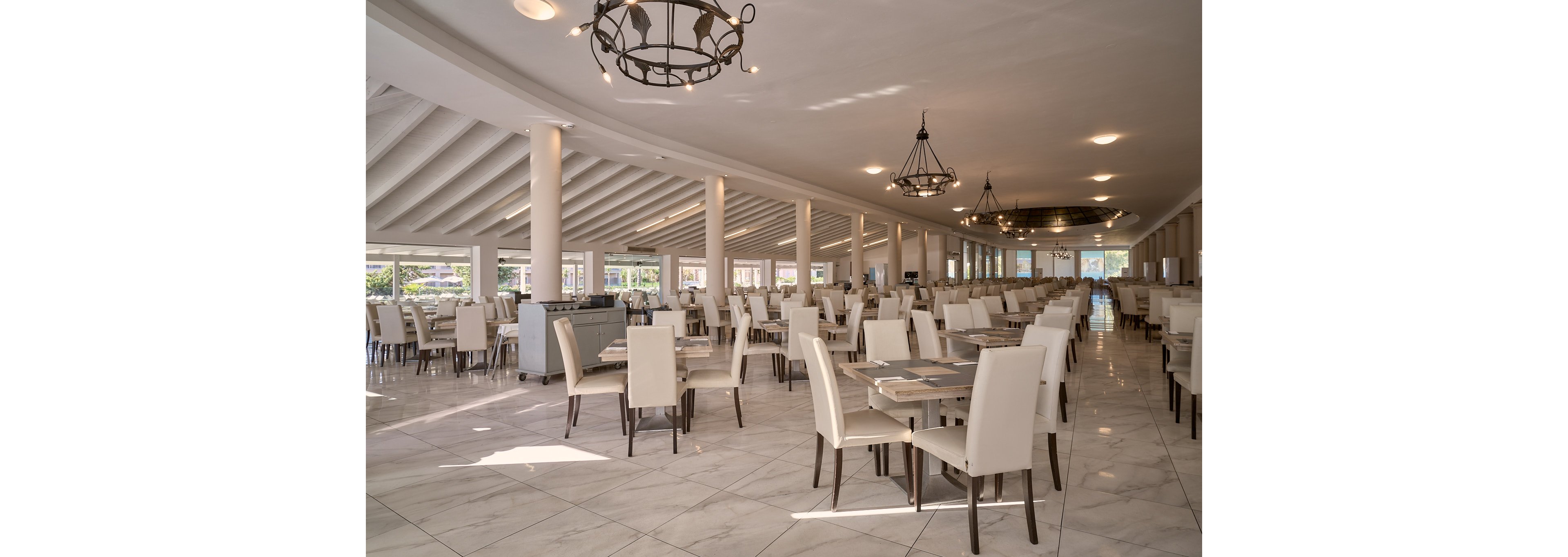 Large, bright restaurant dining hall with rows of neatly arranged tables and cream-colored chairs. The space features a high, sloped ceiling with exposed beams and multiple decorative chandeliers. Sunlight streams in through wide windows, revealing an outdoor view beyond the seating area. The polished tile floor and neutral color palette create a clean, modern, and welcoming atmosphere.