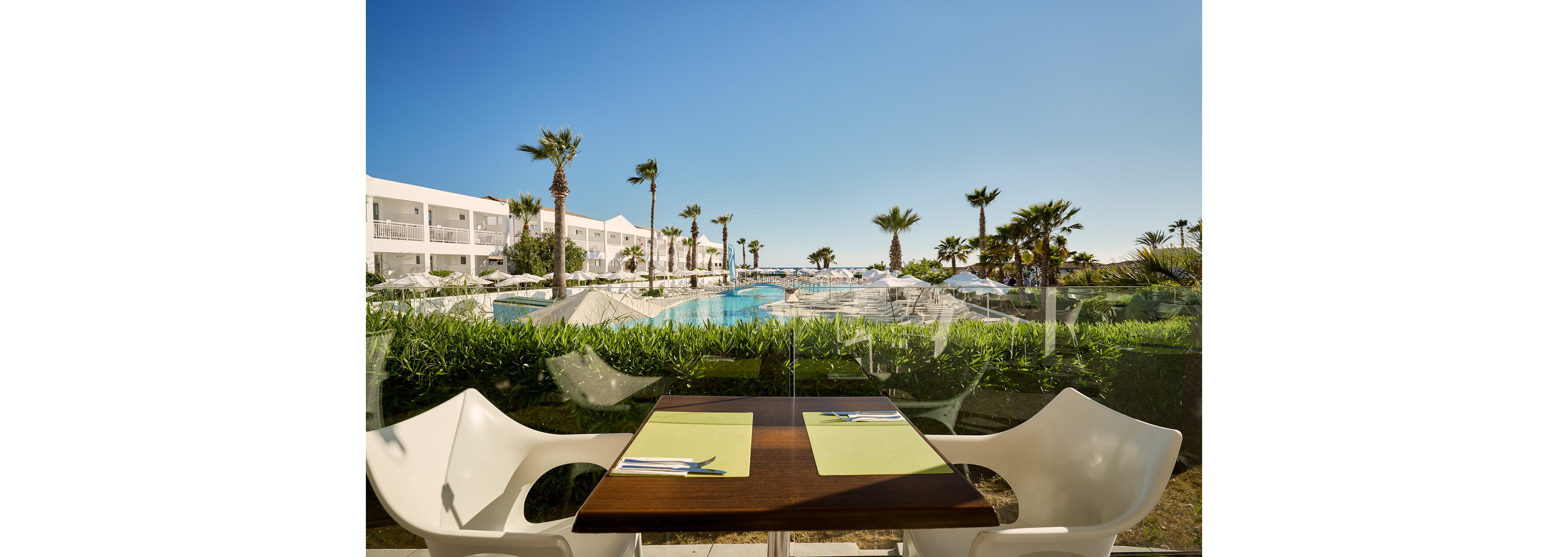 A modern outdoor dining table is set for two overlooking a large resort swimming pool. White chairs and green placemats create a clean, contemporary look in the foreground. Beyond the glass barrier, sun loungers, umbrellas, and palm trees line the pool area under a clear blue sky. A white hotel building frames the scene, suggesting a relaxing vacation atmosphere by the water.