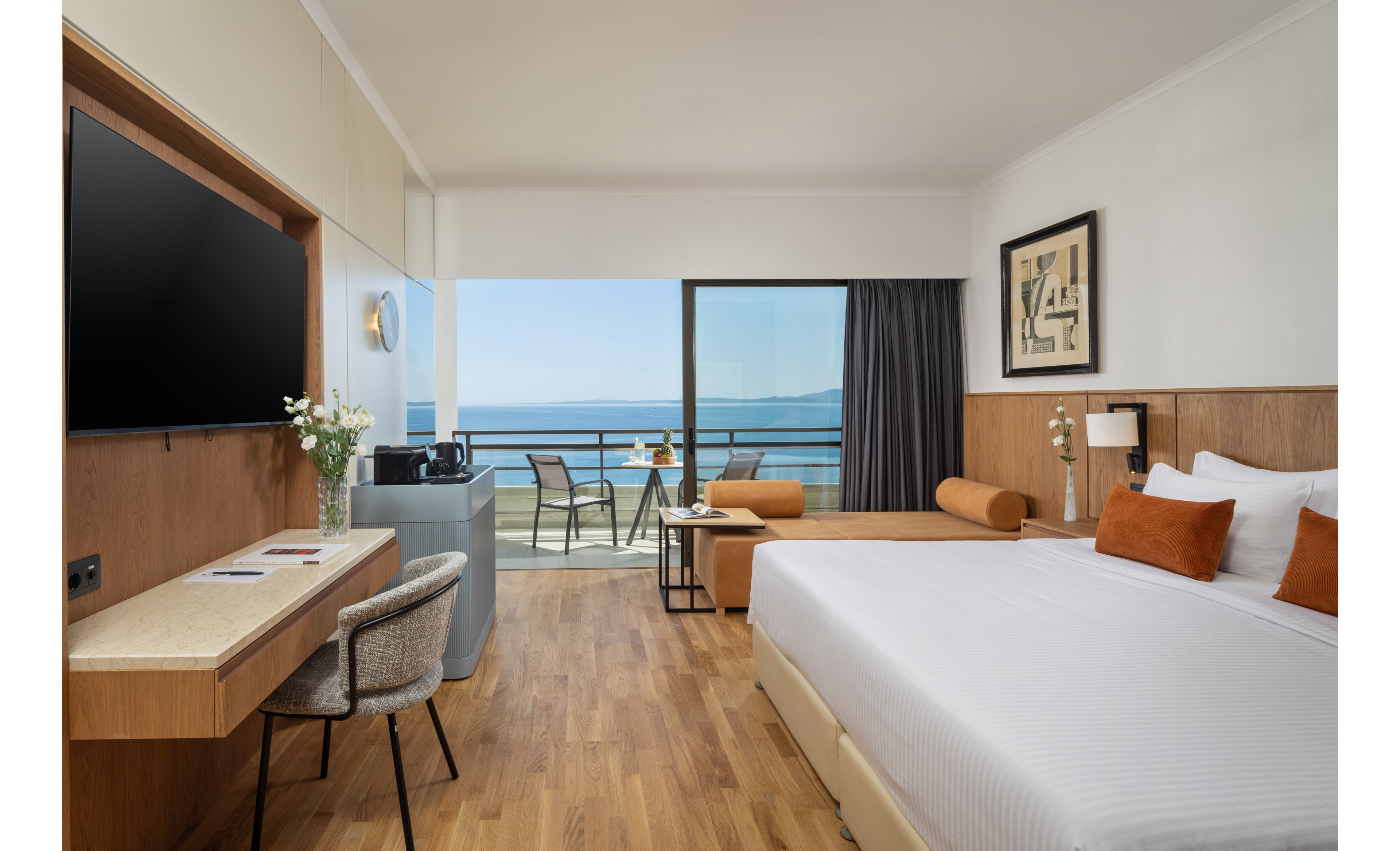 A contemporary hotel room features a large bed with white linens and orange accent pillows. The space includes a wall-mounted television, a minimalist desk with a chair, and a small seating bench. Floor-to-ceiling glass doors open onto a balcony with two chairs and a table overlooking the ocean. Warm wood flooring and neutral walls create a clean, relaxing atmosphere.