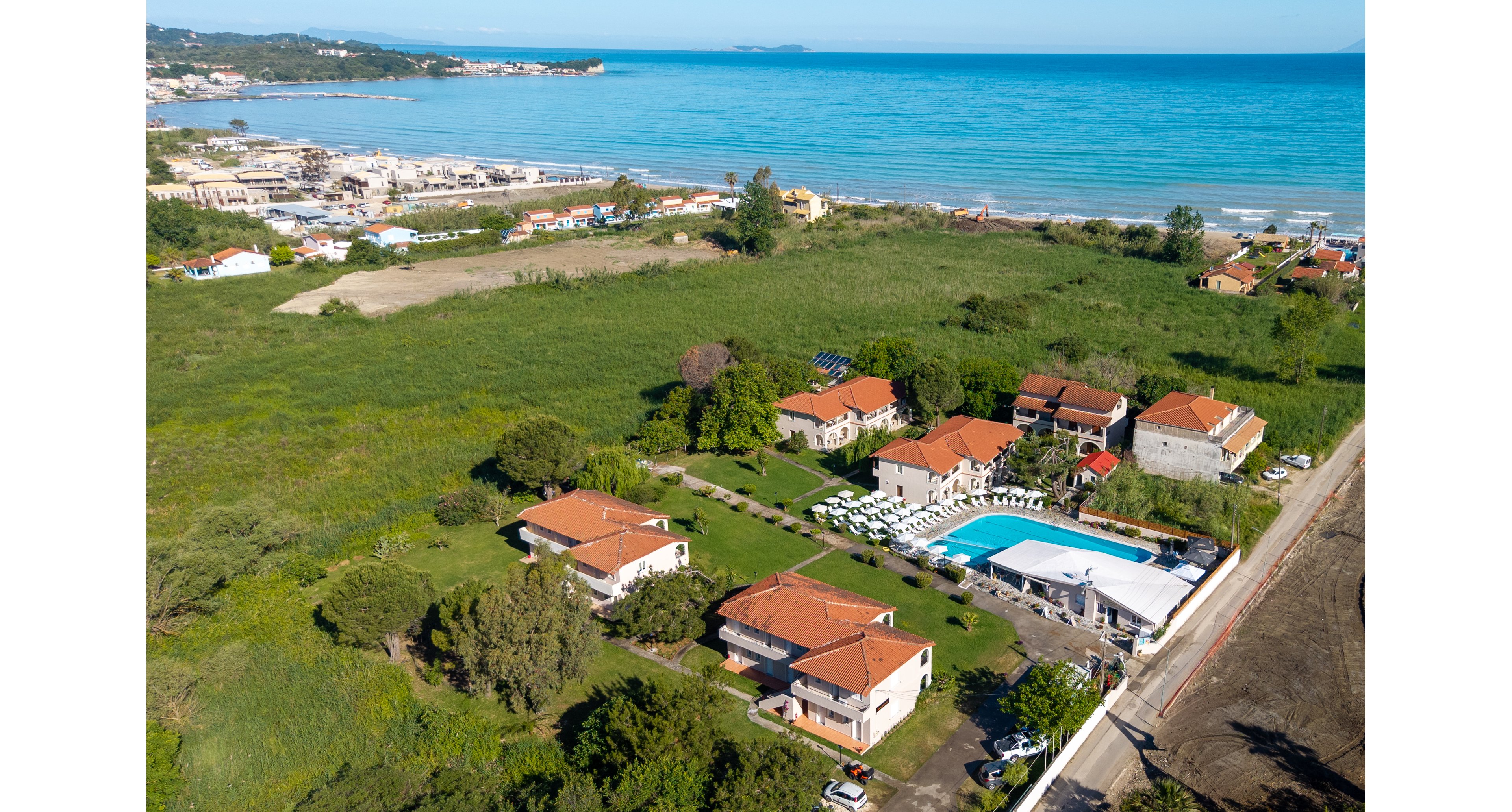 A seaside resort complex with multiple red-roofed buildings is captured from above near a blue ocean shoreline. The property features a large outdoor swimming pool surrounded by sun loungers and umbrellas. Green fields and trees extend around the resort, with additional coastal development visible in the distance. The scene conveys a relaxed holiday atmosphere in a sunny coastal setting.