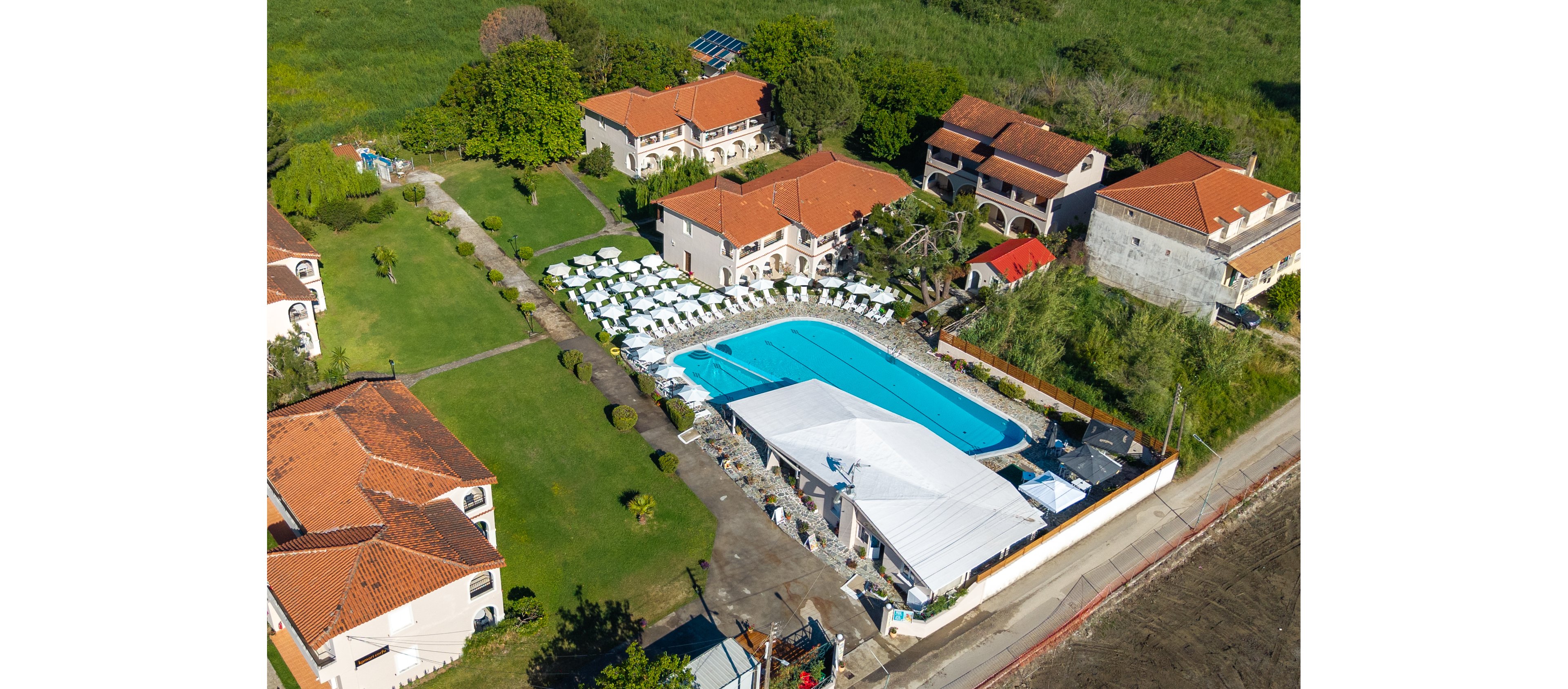 A large outdoor swimming pool is surrounded by white sun loungers and umbrellas in the center of a resort complex. Several low-rise villas with red-tiled roofs are arranged around the pool, separated by green lawns and pathways. A white canopy structure covers part of the poolside area, suggesting a shaded lounge or event space. The scene is set in a lush, green environment at the edge of a more rural landscape, captured from an elevated aerial perspective.