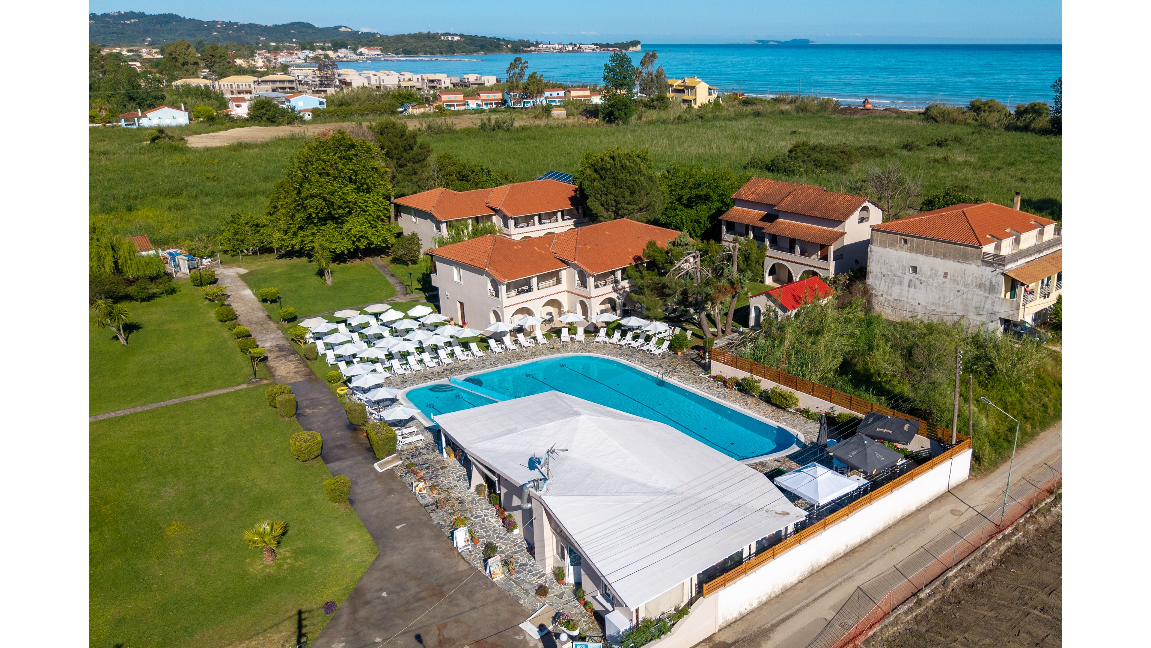 A seaside resort complex features a large central swimming pool surrounded by white sun loungers and umbrellas. Multiple low-rise buildings with red-tiled roofs are arranged around the pool area, with pathways connecting the structures. Green lawns and landscaped gardens extend toward the edge of the property, while the blue sea and distant coastline are visible in the background. The scene is captured from an elevated viewpoint, highlighting the resort layout and its proximity to the beach.