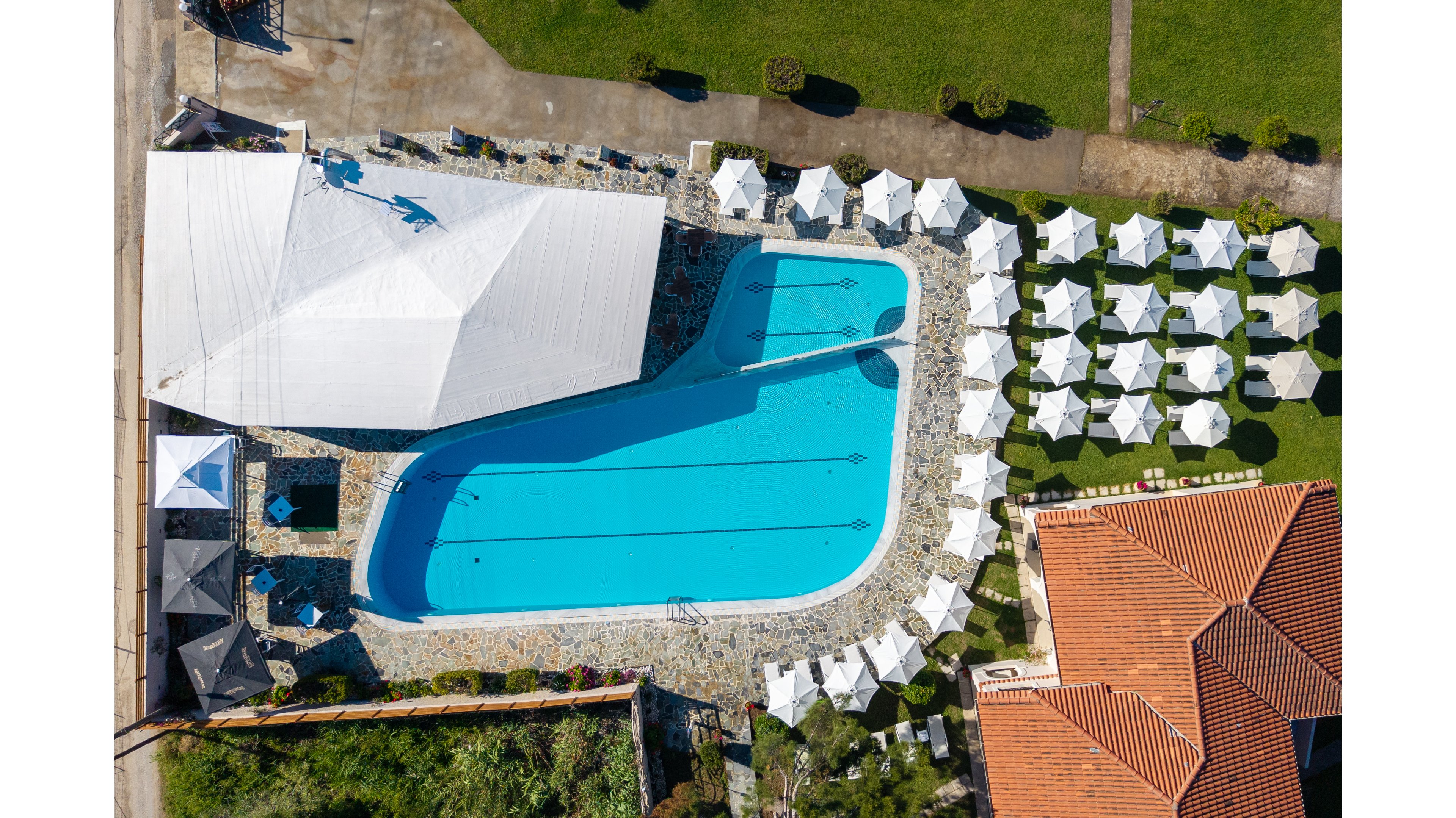 A large outdoor swimming pool is captured from above within a resort or hotel complex. The pool is surrounded by stone decking and a series of neatly arranged white sun umbrellas and lounge chairs. A large white canopy structure provides shaded seating on one side, while red-tiled roofs and green lawns frame the scene. The bright blue water and orderly layout create a clean, inviting vacation atmosphere.