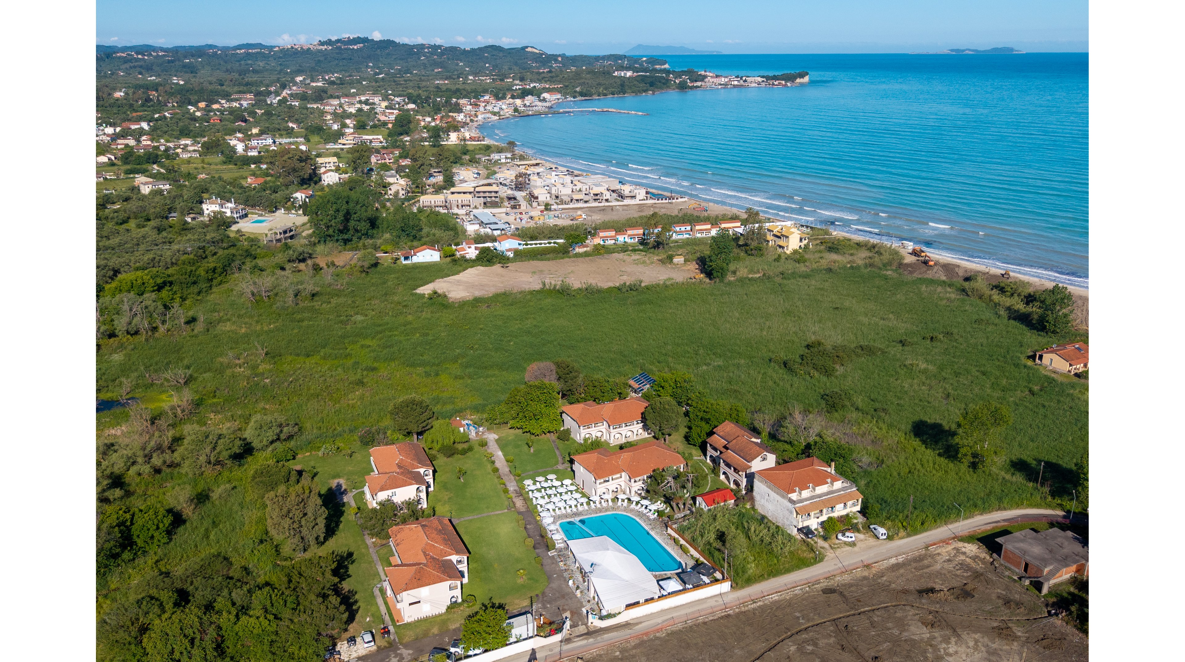 A seaside resort complex with several red-roofed buildings and a central swimming pool is captured from above. The property is surrounded by green fields and vegetation, with a sandy beach and calm blue sea in the background. Sun loungers and umbrellas are arranged around the pool, suggesting a vacation or leisure setting. A coastal town stretches along the shoreline, framed by distant hills under clear skies.