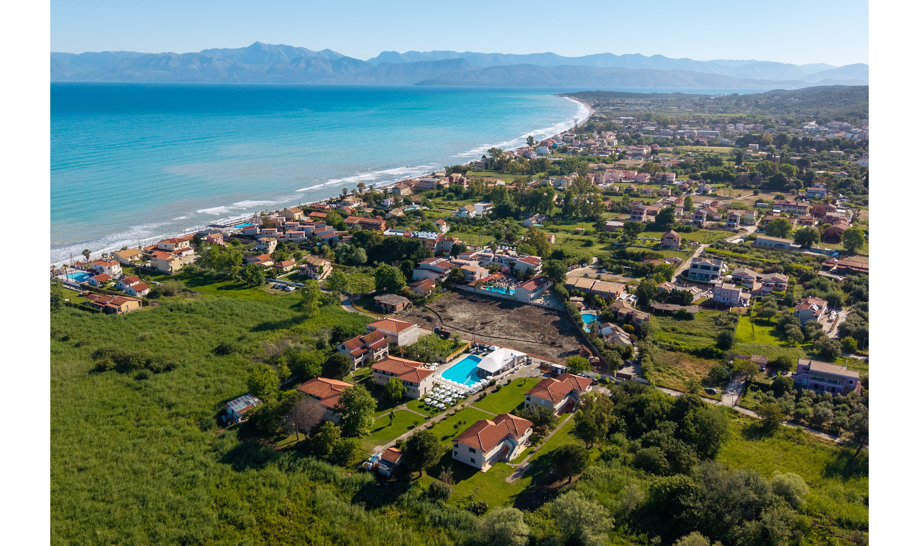 Aerial view of a coastal village stretches along a bright turquoise shoreline. Houses with red-tiled roofs and small roads are surrounded by lush green vegetation. A central resort complex features a long rectangular swimming pool and manicured lawns. In the distance, mountains rise above the horizon, framing the expansive beach and seaside landscape.