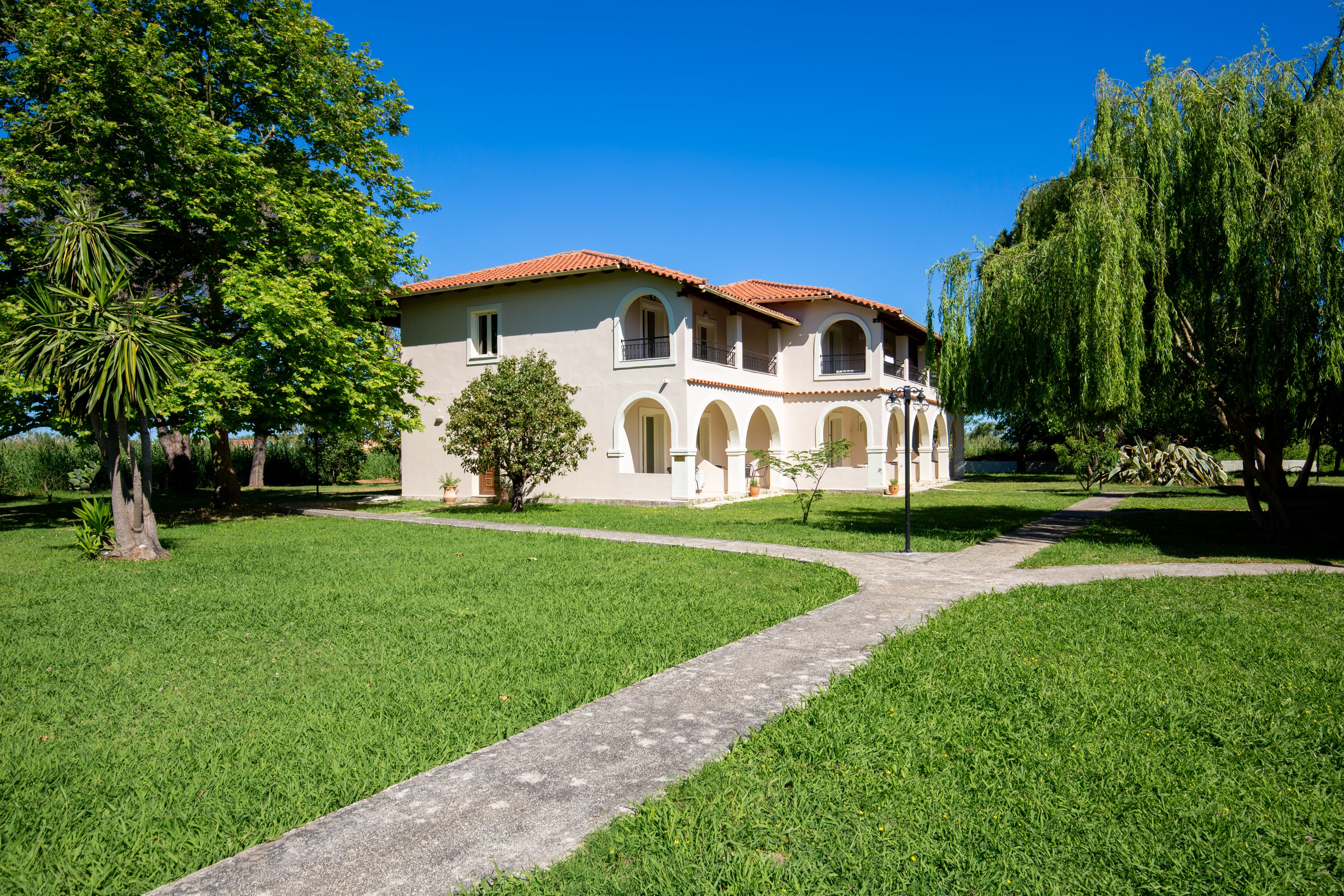A two-story cream-colored villa with a red tiled roof stands in the middle of a lush green lawn. Arched balconies and doorways give the house a Mediterranean architectural style. A concrete path leads to the entrance, surrounded by trees and manicured grass under a clear blue sky. The bright daylight and vivid colors create a peaceful, inviting atmosphere.