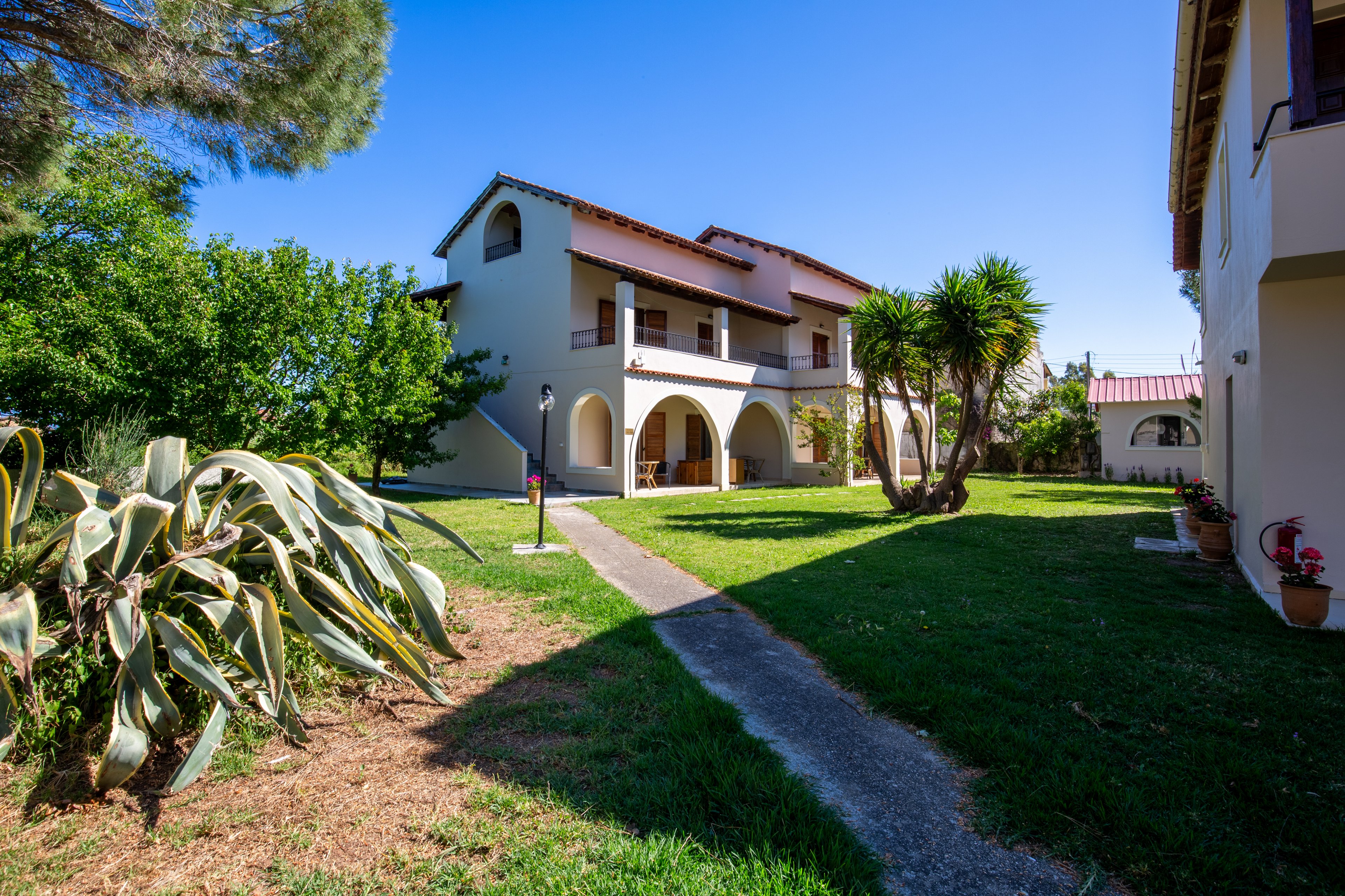 A two-story Mediterranean-style house with white walls and terracotta roof tiles stands in a bright, sunny yard. A narrow concrete path leads from the foreground toward the arched front porch and entrance. Lush green grass, trees, and large succulents frame the building, creating a peaceful residential setting. Another similar building is visible to the right, suggesting a small housing complex or villa property.