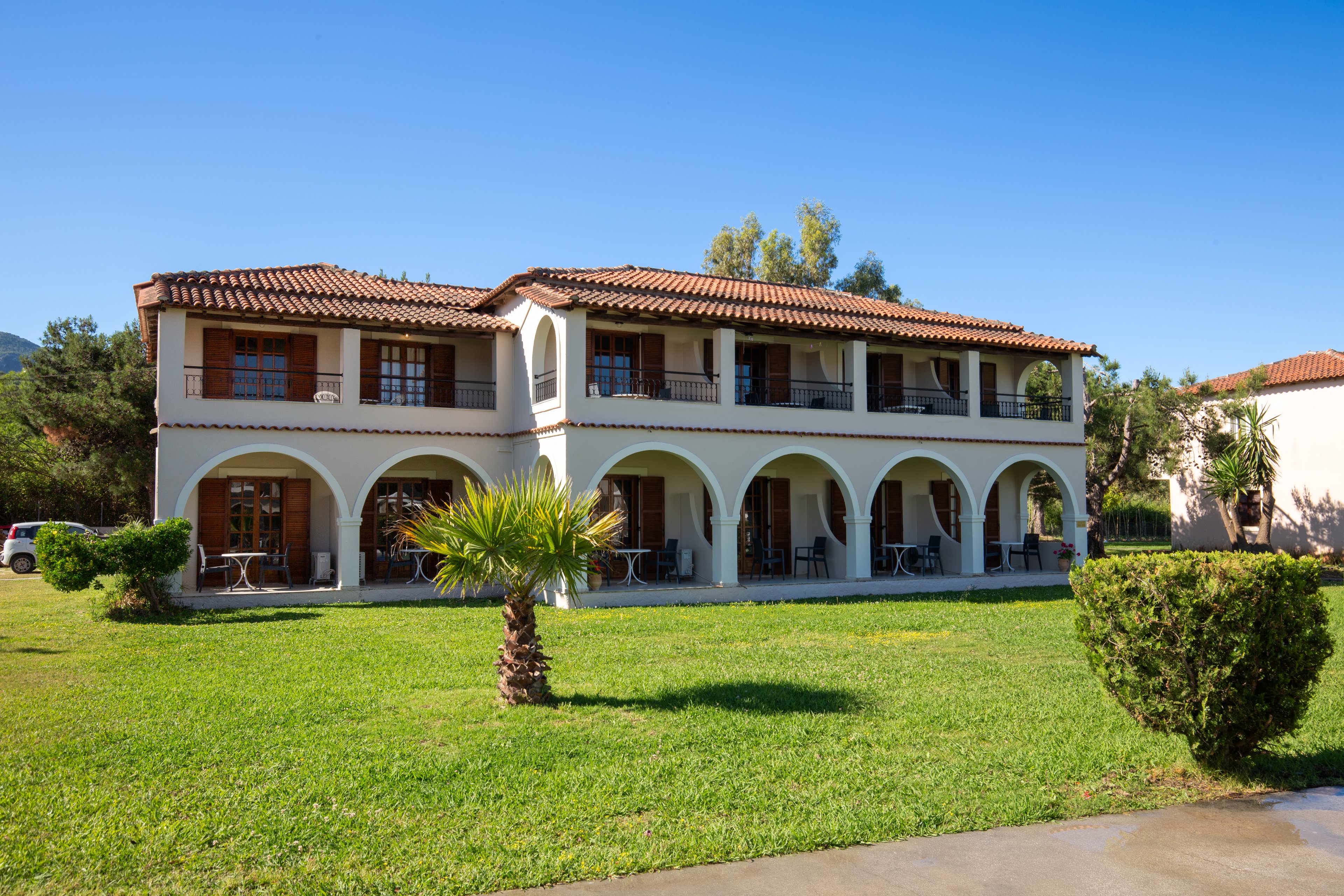 Two-story Mediterranean style building stands in a sunny outdoor setting. The structure features white walls, terracotta roof tiles, and a series of arches on the ground floor. Balconies with railings and wooden shutters add detail to the upper level. A well-maintained green lawn, small palm tree, and trimmed bushes surround the villa under a clear blue sky.