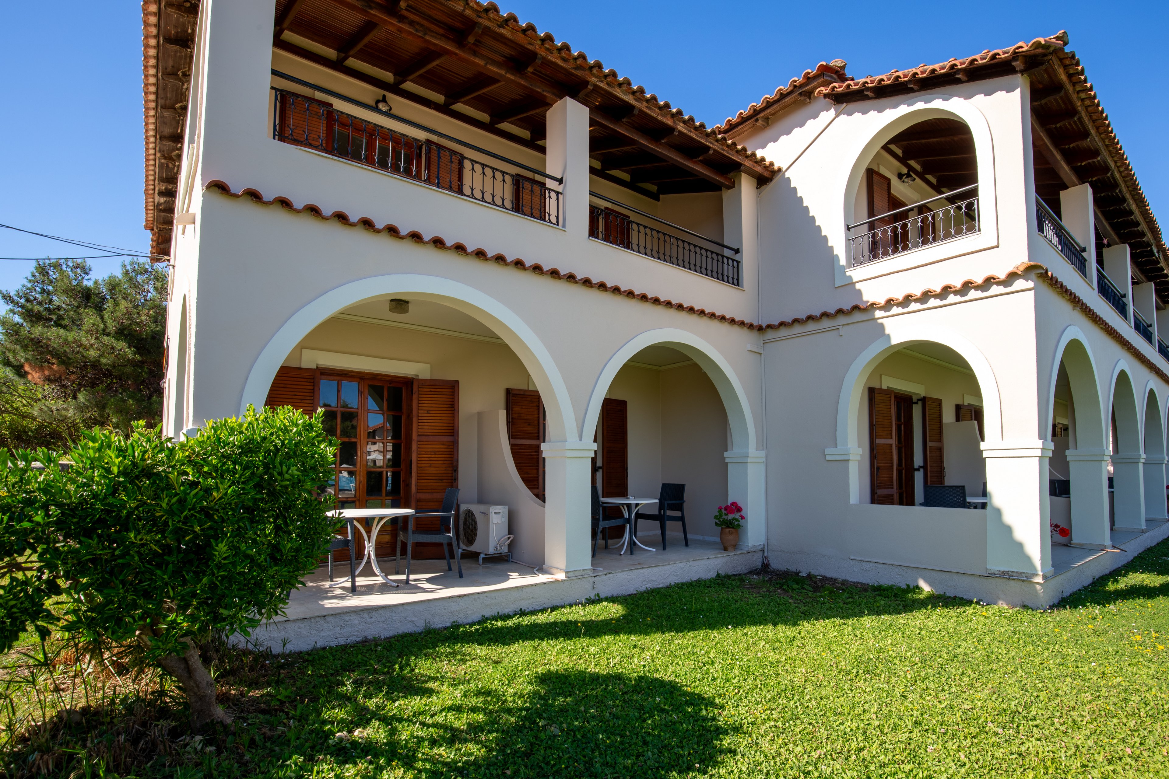 Two-story cream-colored villa features prominent arched verandas and wooden shutters. The house is set in a sunny outdoor environment with a neatly maintained green lawn in the foreground. Patio tables and chairs are placed under the arches, suggesting a relaxing outdoor living space. Clear blue sky and surrounding greenery enhance the warm, inviting atmosphere of the property.