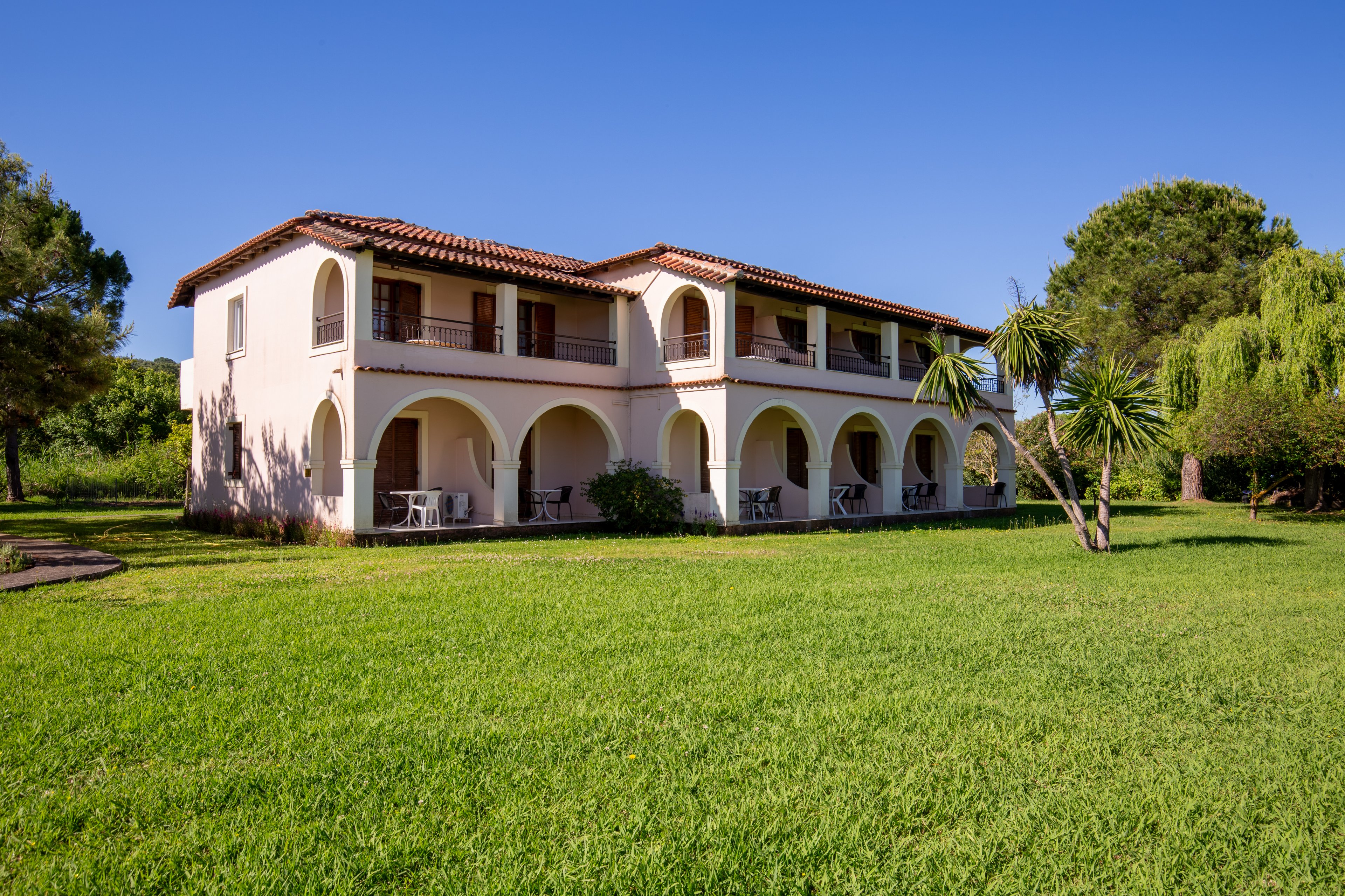 A two-story Mediterranean-style villa stands in the middle of a lush green lawn. The building features white walls, terracotta roof tiles, and a series of large arches forming covered terraces on both levels. Outdoor chairs and tables are visible under the arches, suggesting a relaxing residential or vacation setting. Tall trees and shrubs surround the property under a clear blue sky, creating a peaceful countryside atmosphere.