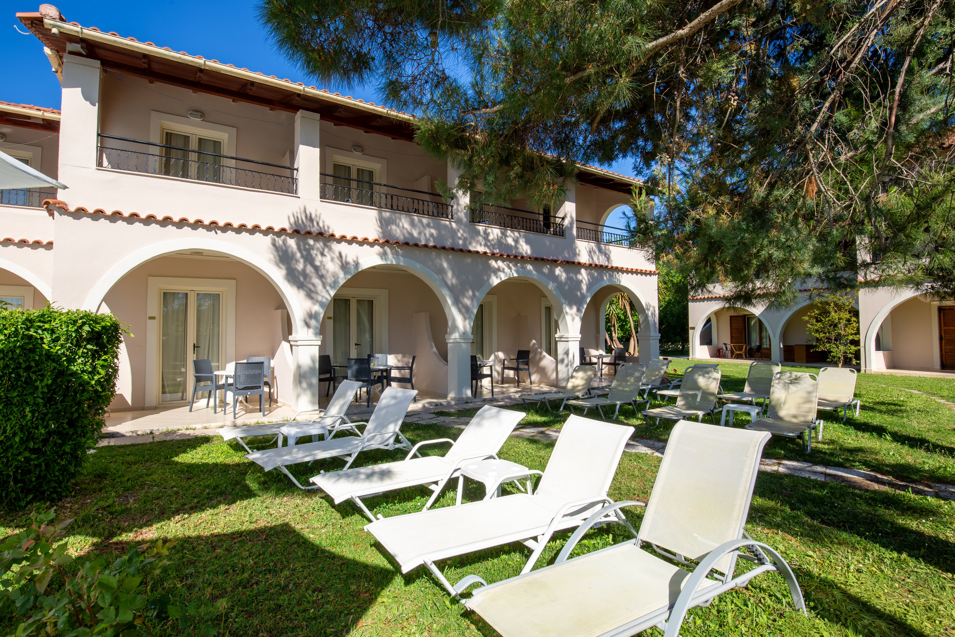 A two-story light-colored hotel building features arched terraces and balconies overlooking a grassy garden. Several white sun loungers are arranged on the lawn, suggesting a relaxing outdoor area for guests. Patio tables and chairs sit under the arches near glass doors leading to the rooms. Bright sunlight, clear blue sky, and surrounding greenery create a warm, inviting vacation atmosphere.