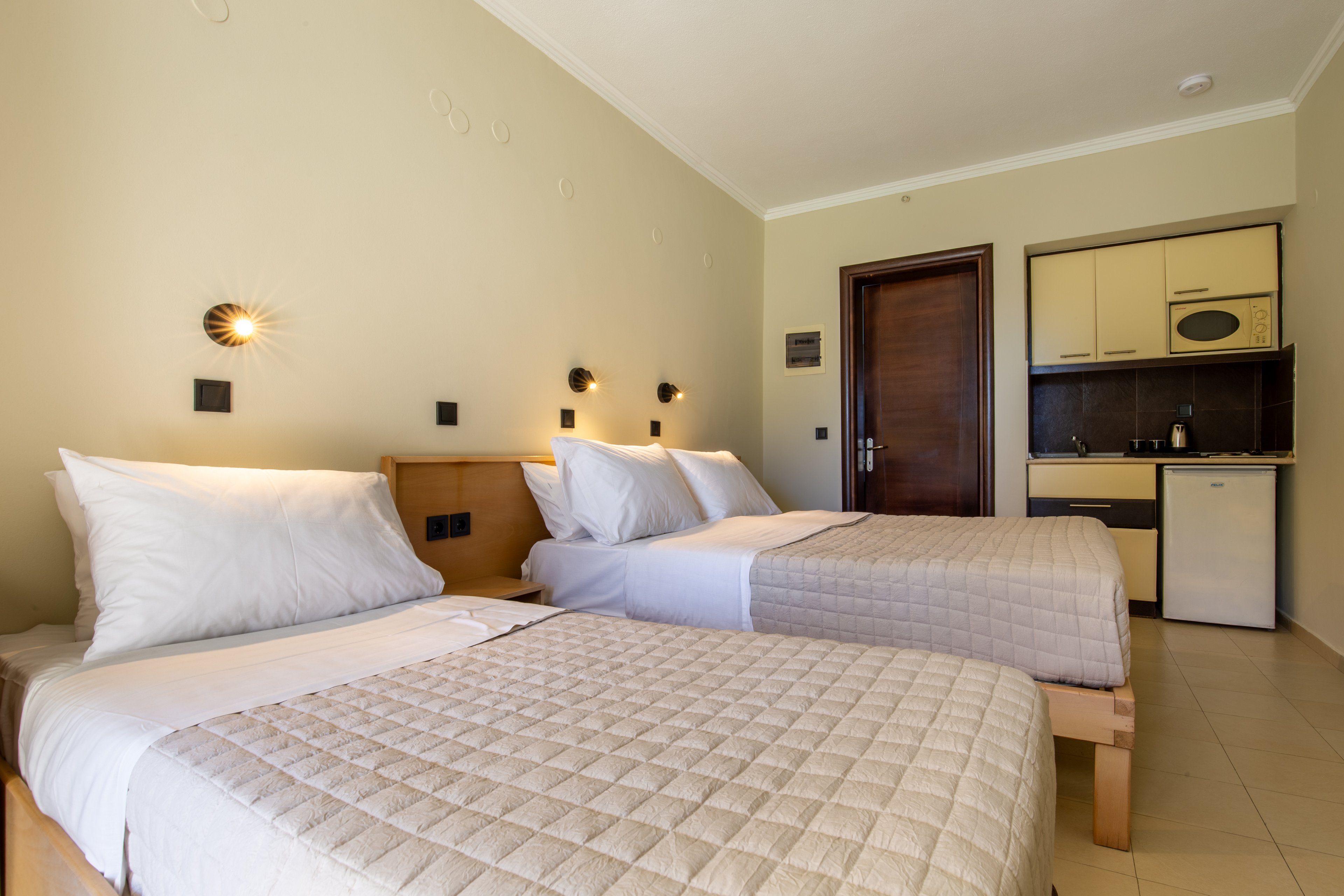 A bright hotel room features two neatly made twin beds with quilted beige bedspreads and white pillows. The space includes a small kitchenette with cabinets, a sink, stovetop, and compact refrigerator along the right wall. Warm wall-mounted lights above the beds create a welcoming atmosphere against the light-colored walls. The tiled floor and simple wooden furniture contribute to a clean, minimalist interior design.
