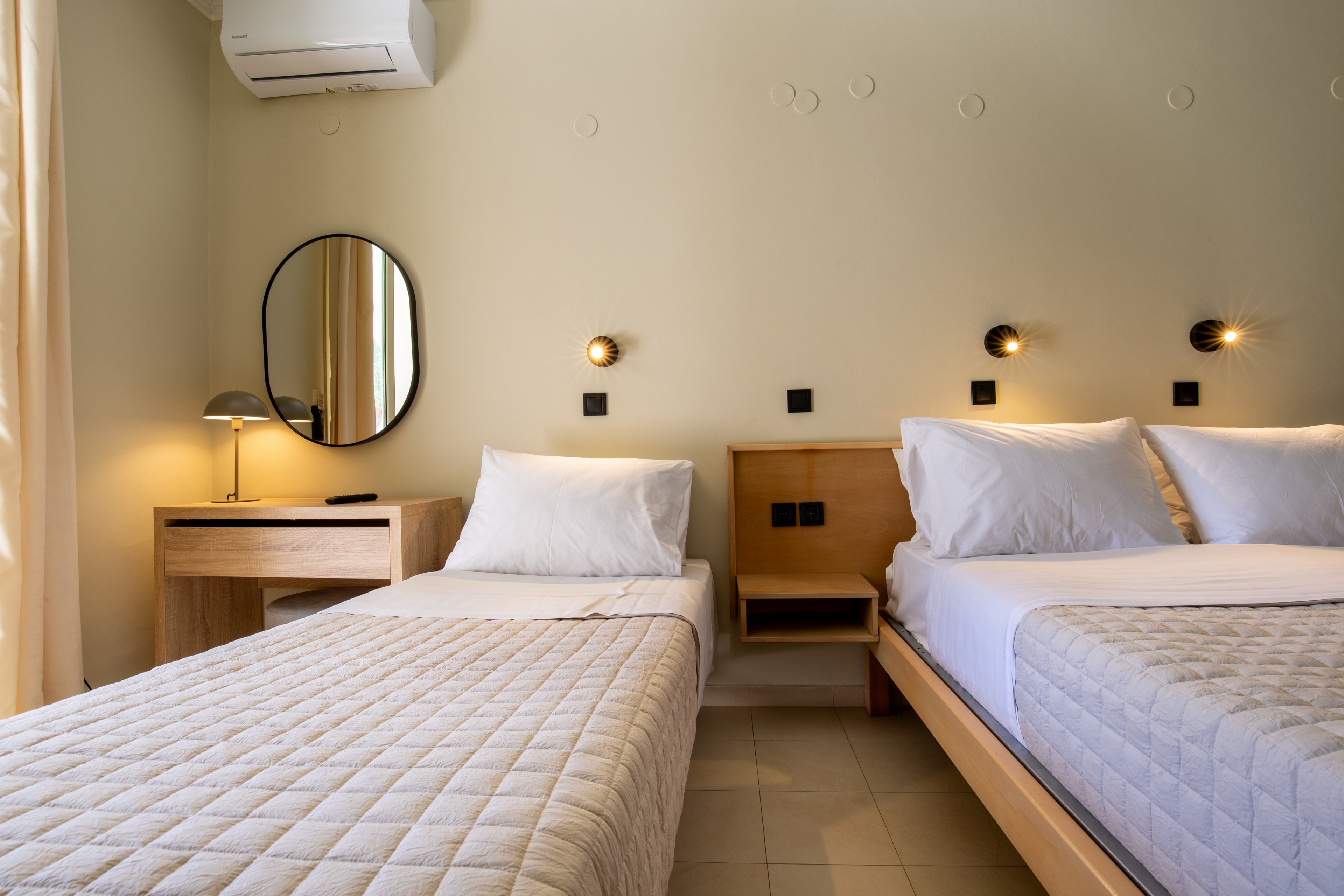 A contemporary bedroom features two neatly made twin beds with quilted bedspreads and white pillows. The room has light-colored walls, tiled flooring, and soft natural light coming through a window with curtains. A wooden desk with a round mirror and table lamp sits beside one bed, while small wall-mounted lights and power outlets are positioned above the headboards. An air conditioning unit is installed high on the wall, contributing to the clean, modern aesthetic.